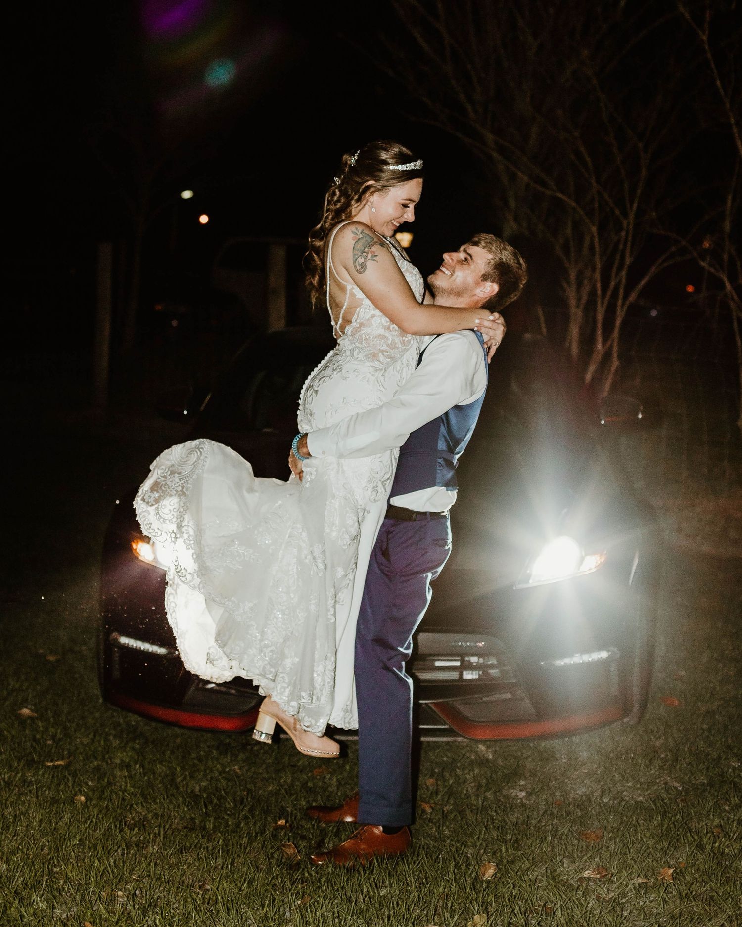 Dramatic nighttime wedding photo with couple embracing against car headlights in background.