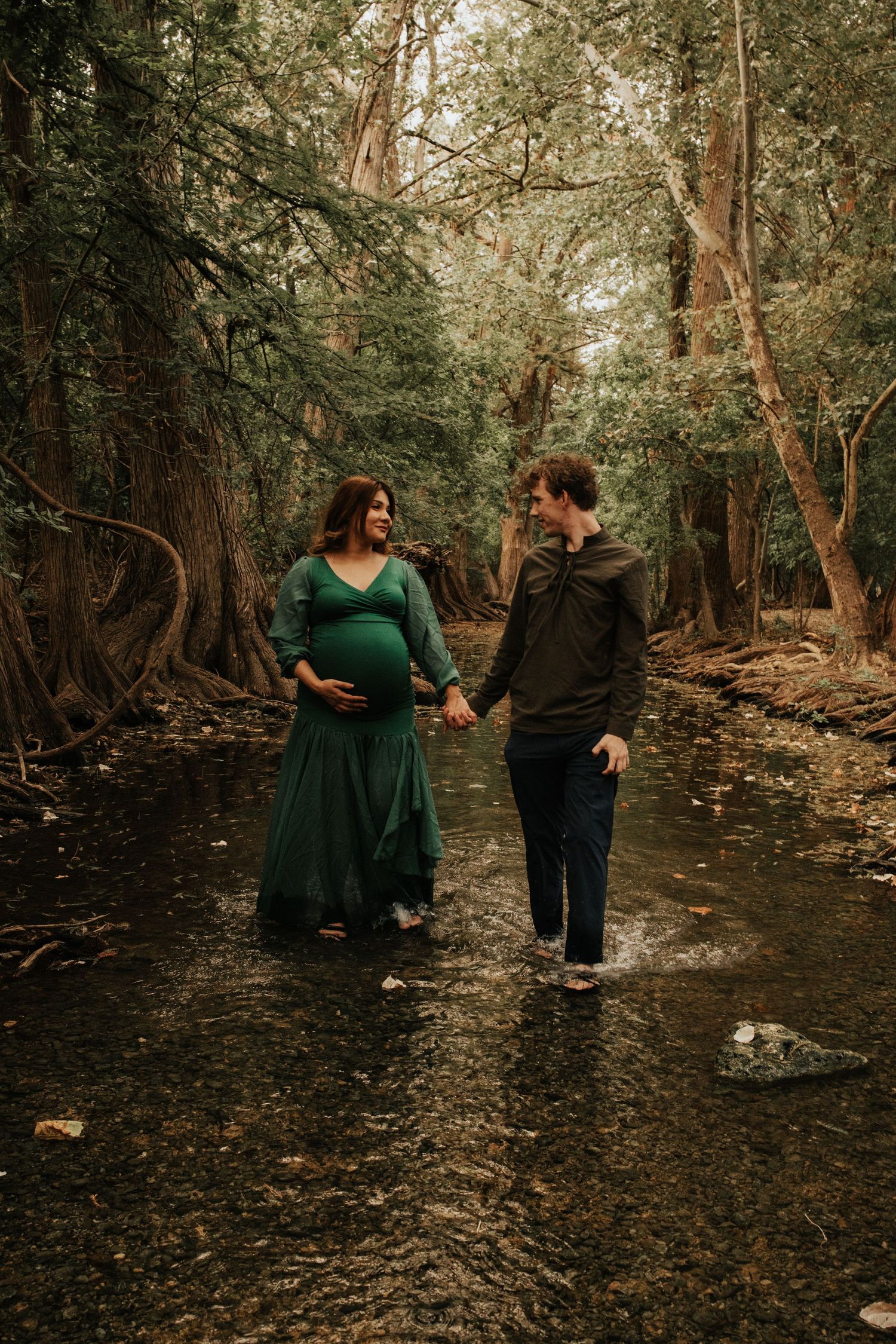 A couple shares a romantic moment while standing in a creek surrounded by lush forest trees and autumn foliage.