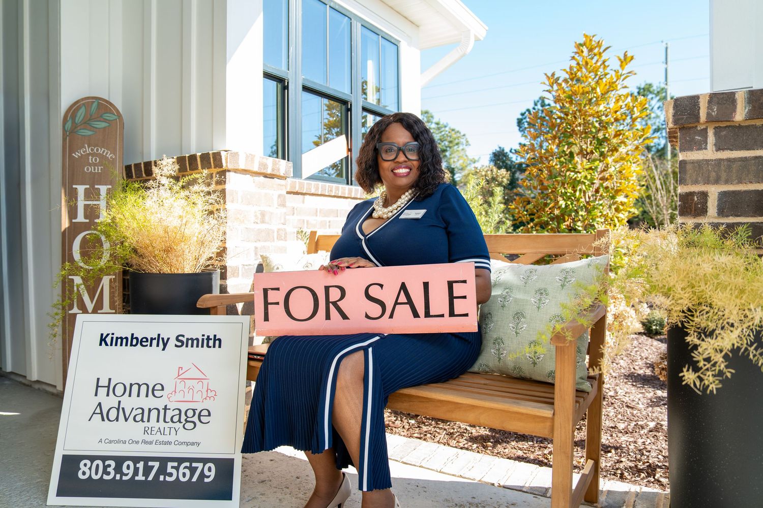 Woman in a navy dress sits on a porch bench holding a For Sale sign beside a Home Advantage Realty yard sign.