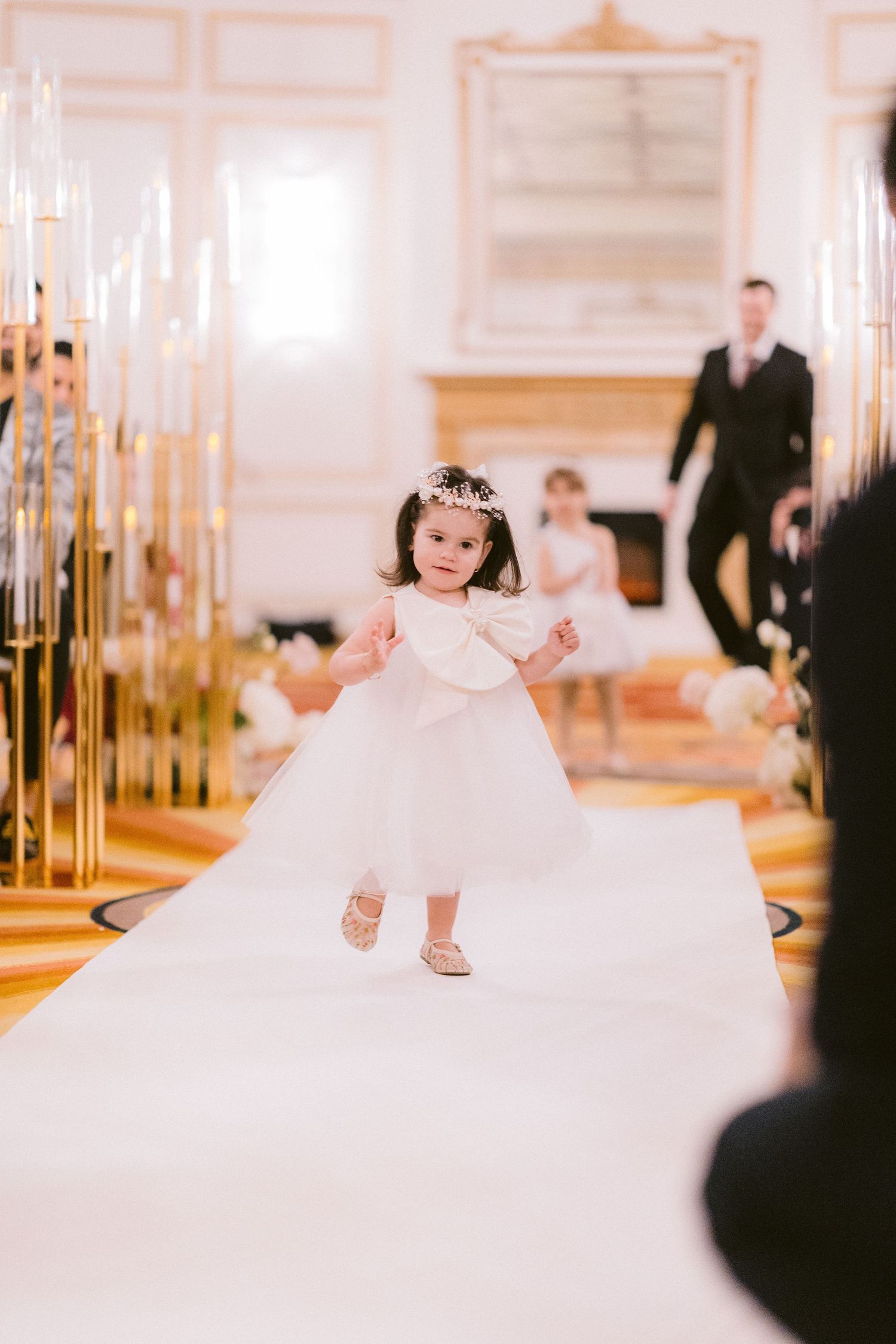 A small child in a white dress and tiara walks down a white aisle lined with tall gold candelabras at an elegant event.