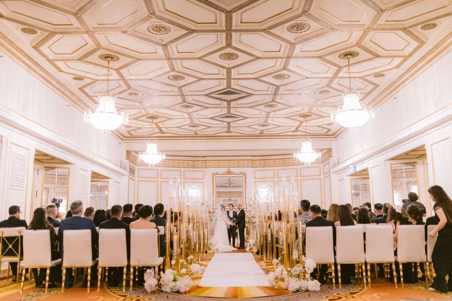 Elegant wedding ceremony in a grand ballroom with ornate ceiling, crystal chandeliers and guests seated along the aisle.