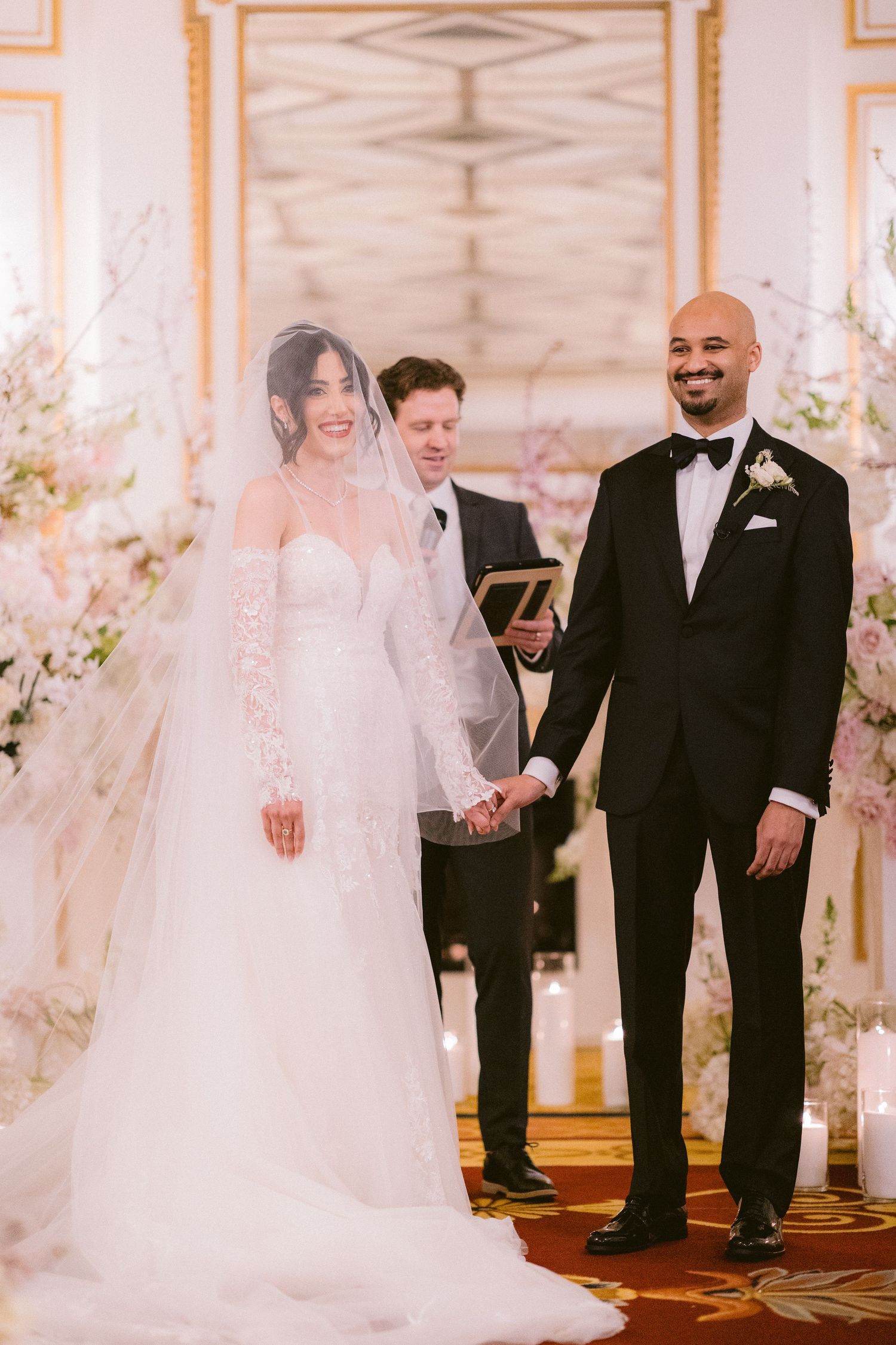 A joyful wedding ceremony taking place in an elegant white and gold ballroom with floral decorations.