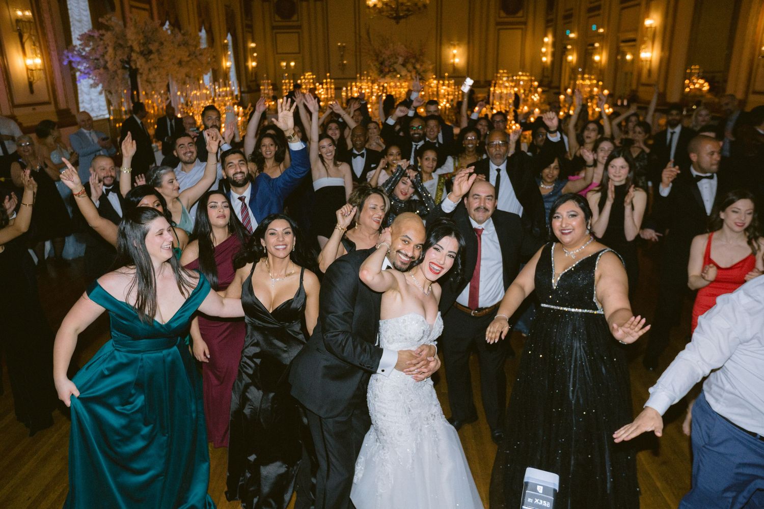 Wedding reception guests dance and celebrate on a crowded dance floor under warm lighting at an evening party.