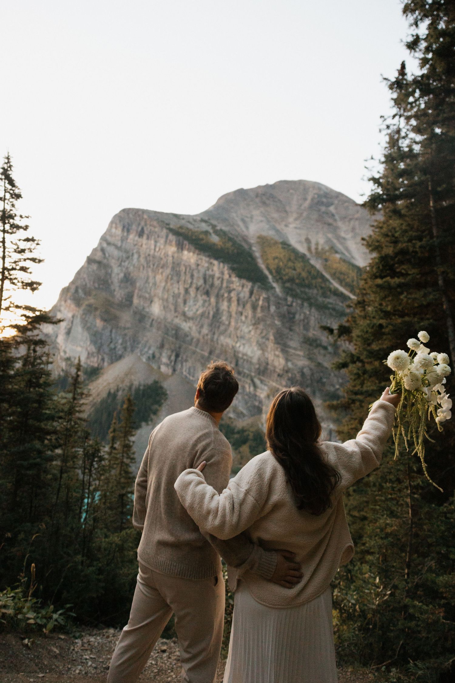 A couple in cozy sweaters embraces while admiring majestic mountain cliffs and evergreen forest scenery.