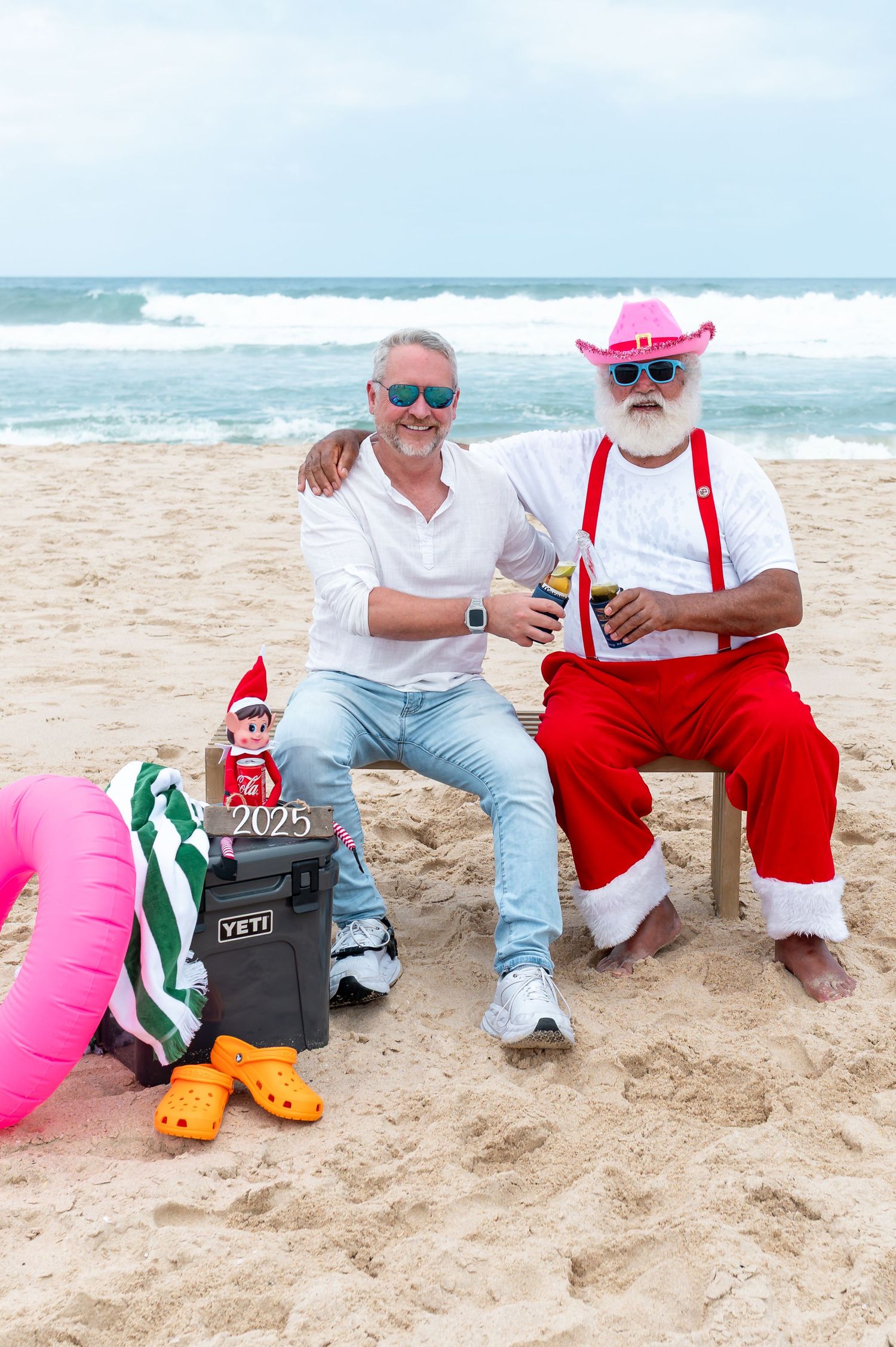 A festive Christmas beach scene with people in Santa hats sitting by decorated driftwood and flowers near the ocean.