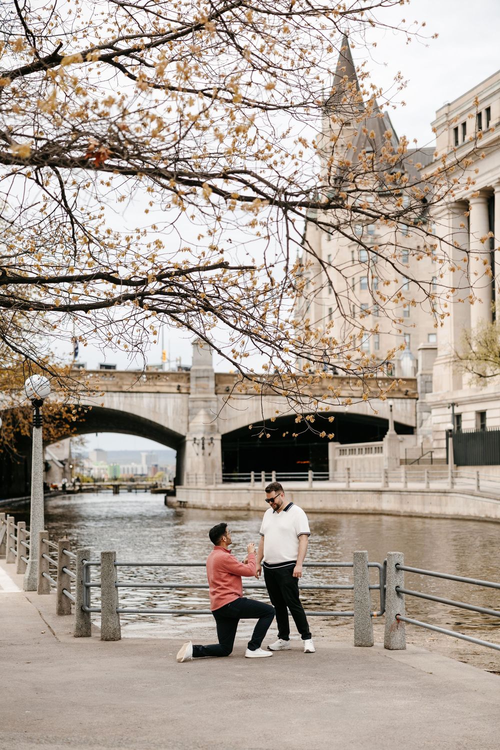 Man proposes to partner on one knee by a river with a historic stone bridge and building in autumn.