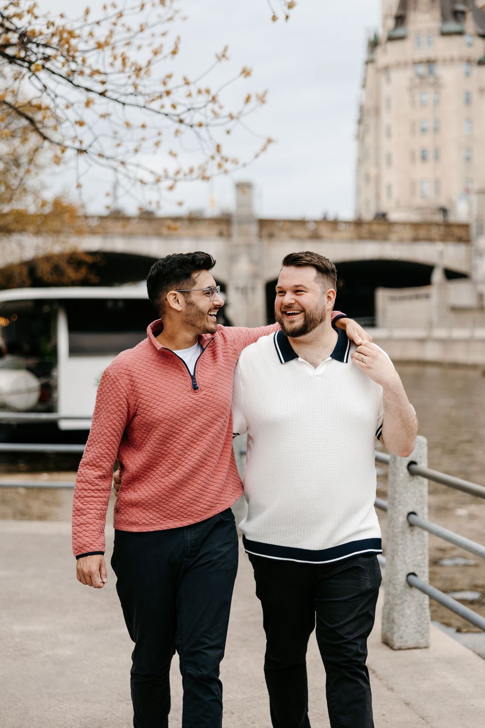 Two men smiling and walking arm in arm along a riverside promenade with a bridge and building in the background.