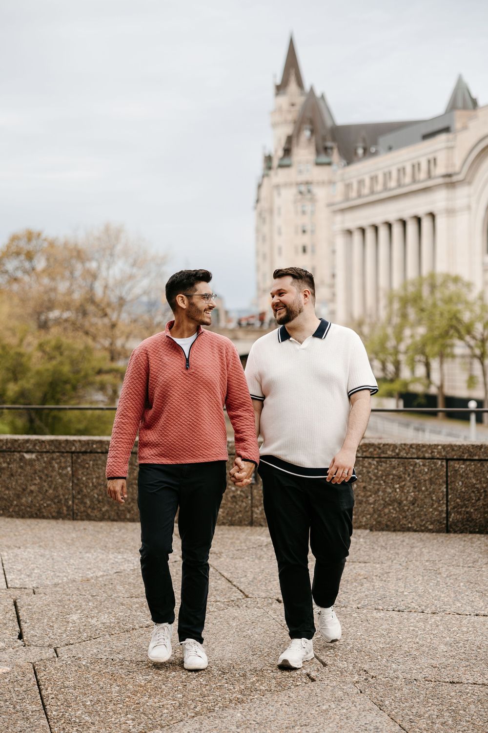 Two men hold hands and smile while walking outdoors near a grand historic building on a cloudy day.