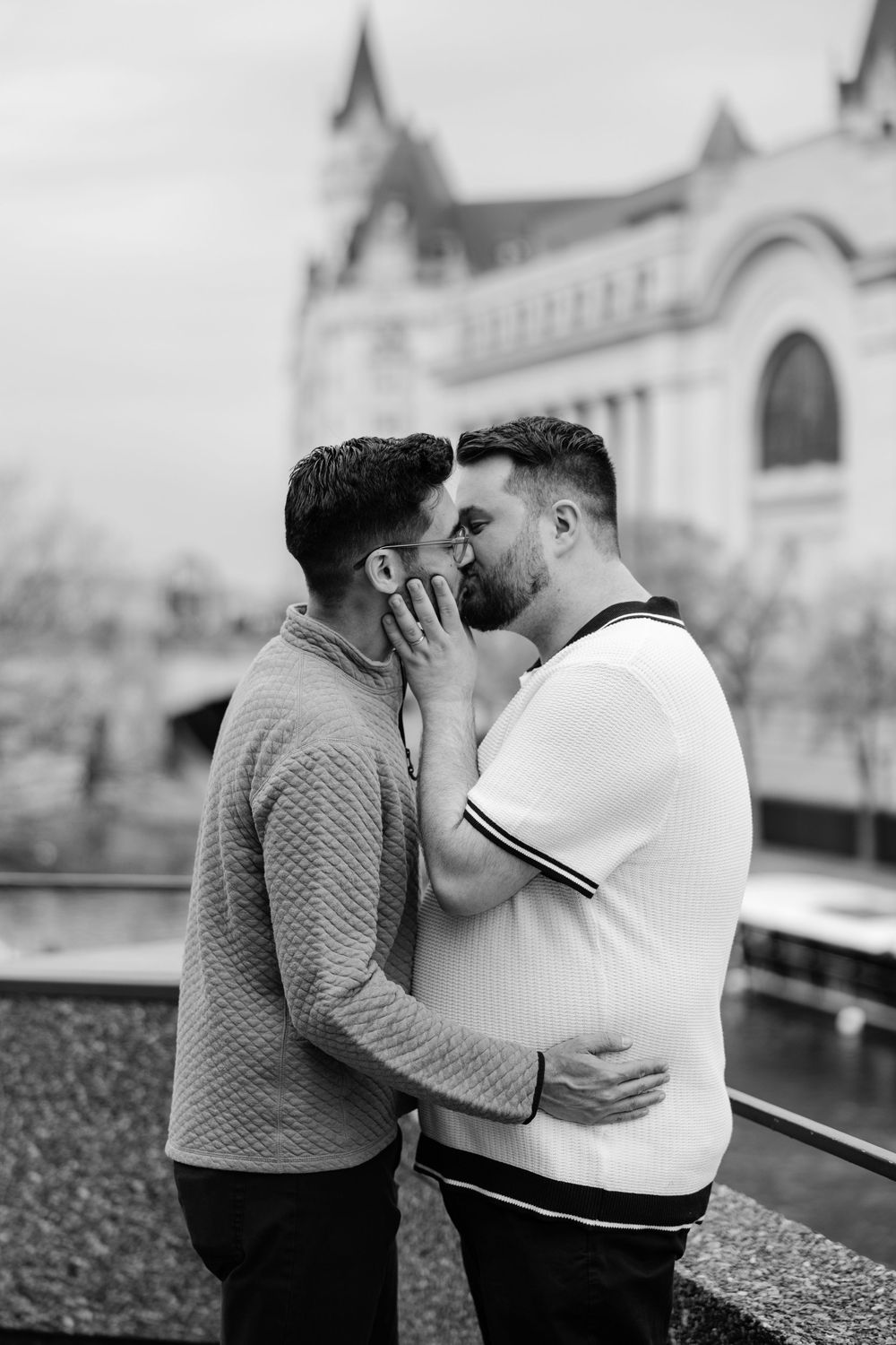 Black and white photo of two men sharing a romantic kiss outdoors with a historic building in the background.