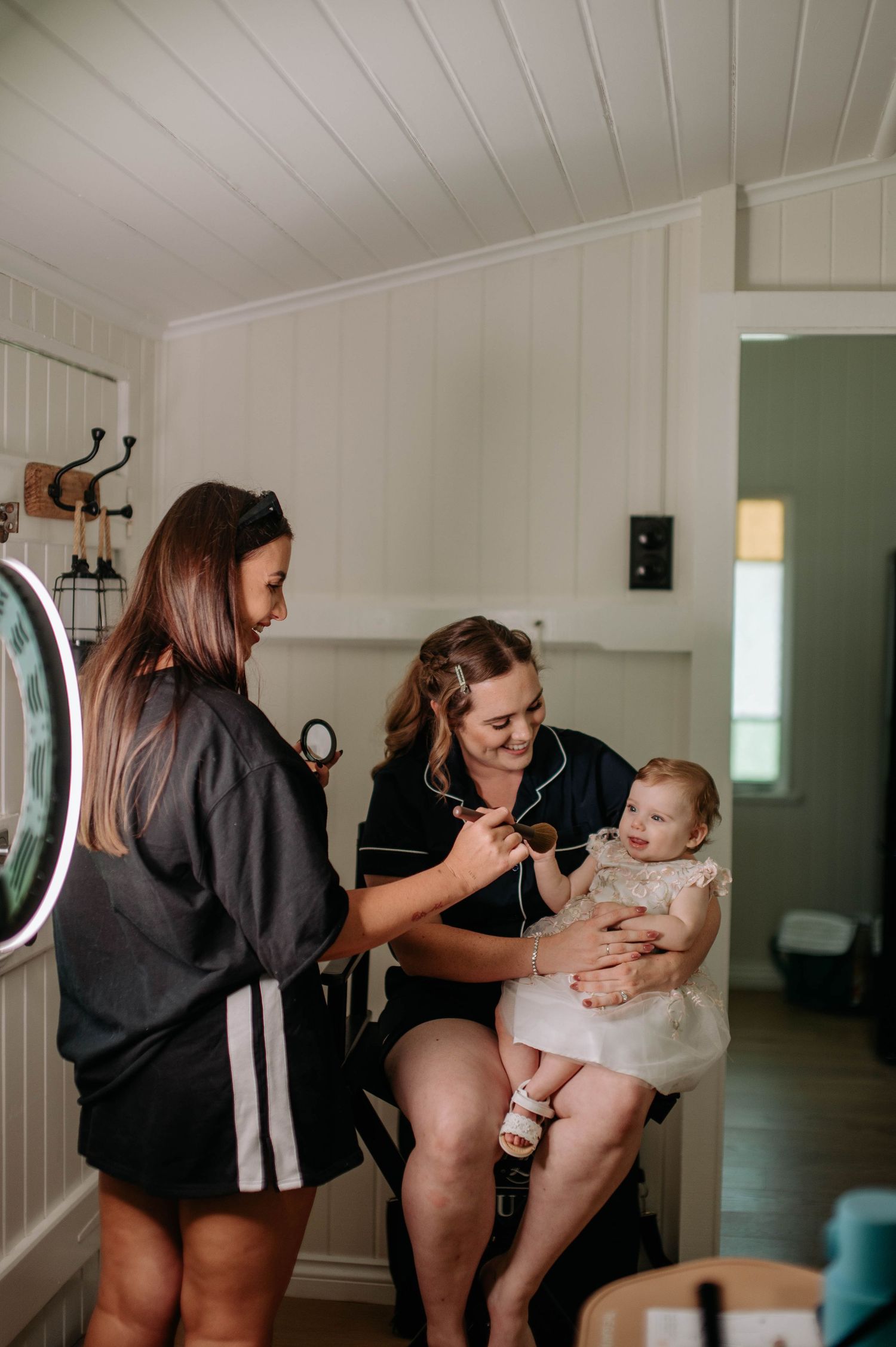 Two people in black outfits interact with a baby wearing a white dress in a bright interior room.