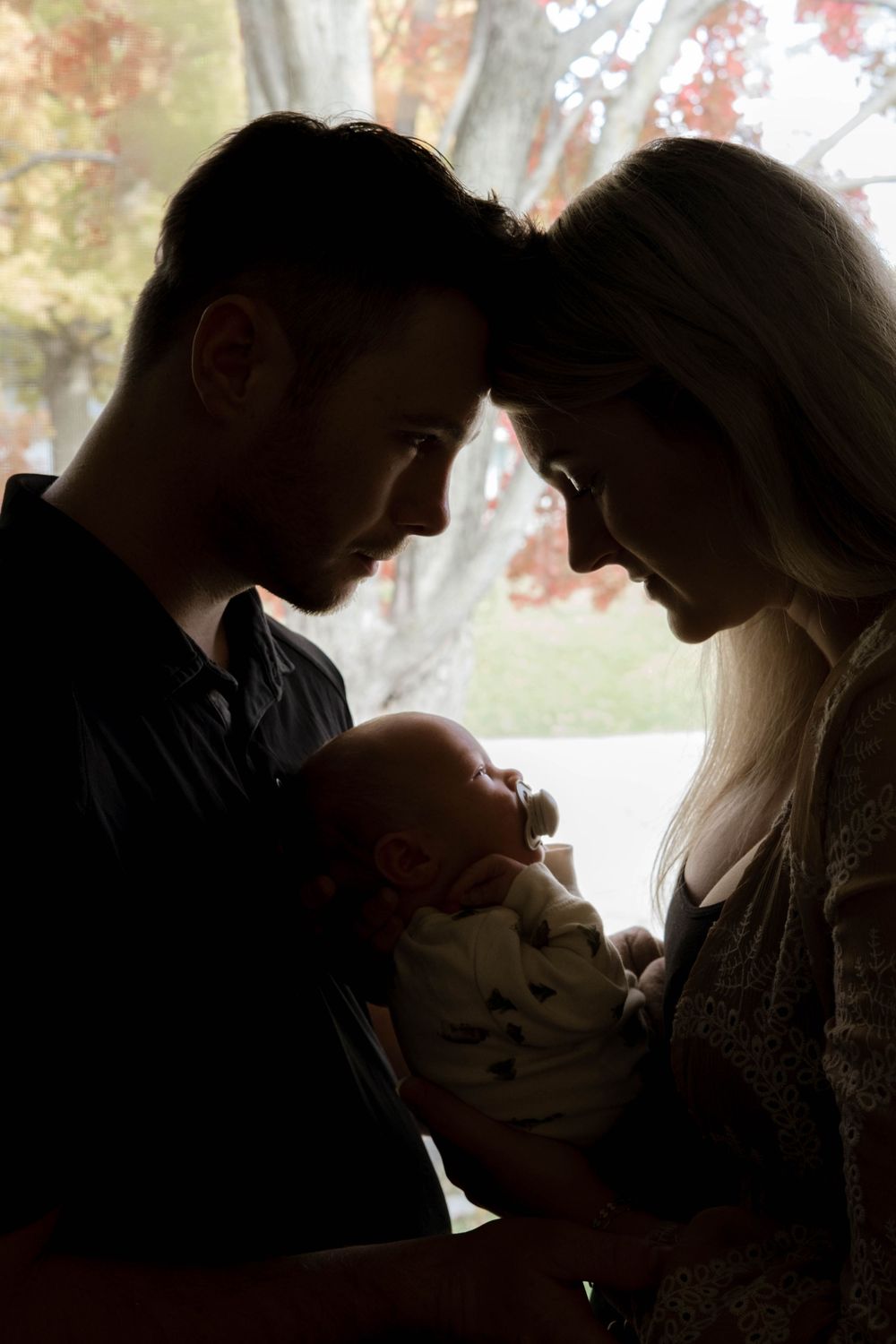 Silhouetted figures share an intimate moment while holding infant by window with autumn leaves.