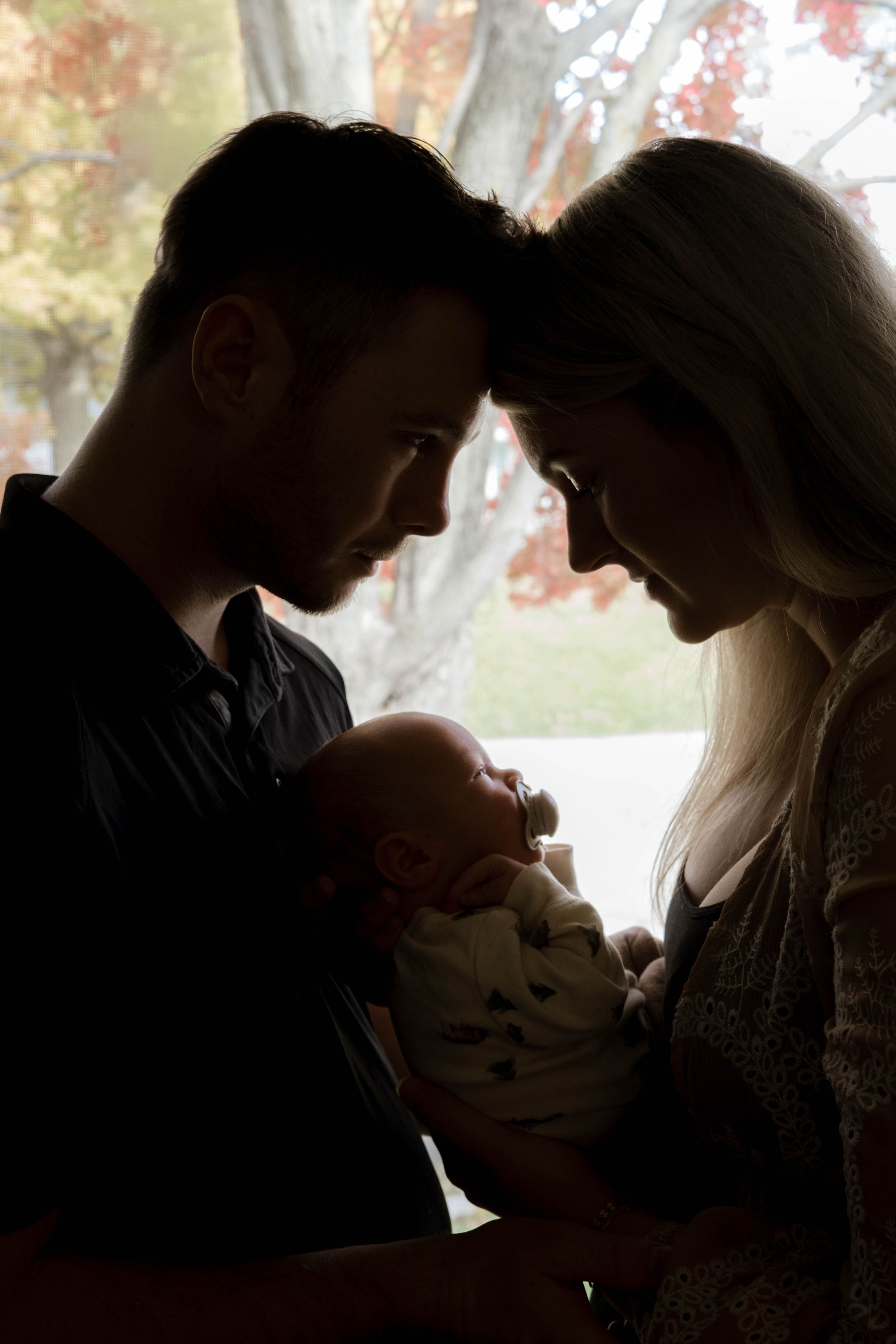 Silhouetted figures share an intimate moment while holding infant by window with autumn leaves.