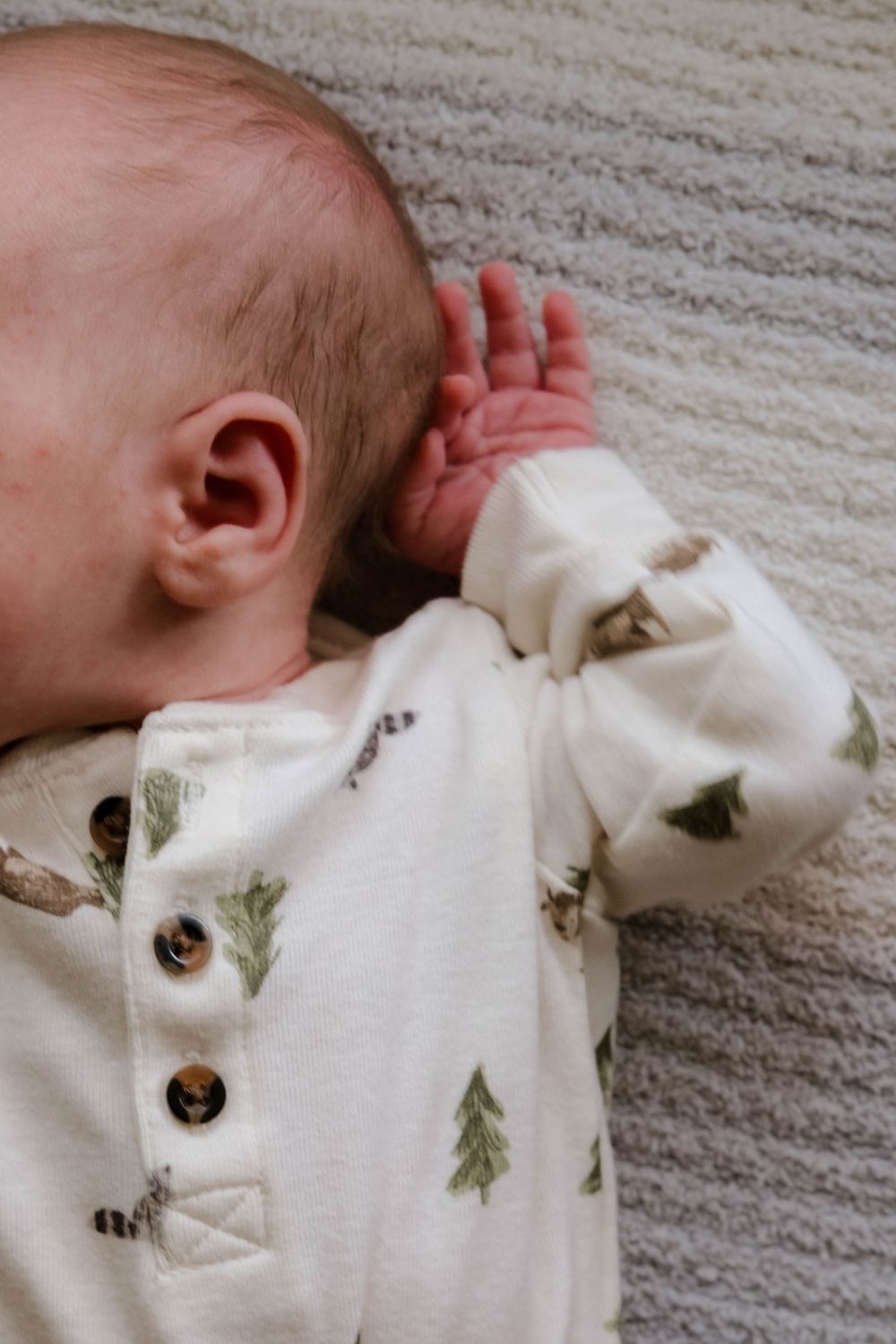 Sleeping infant wearing white onesie with forest pattern lies on textured carpet.