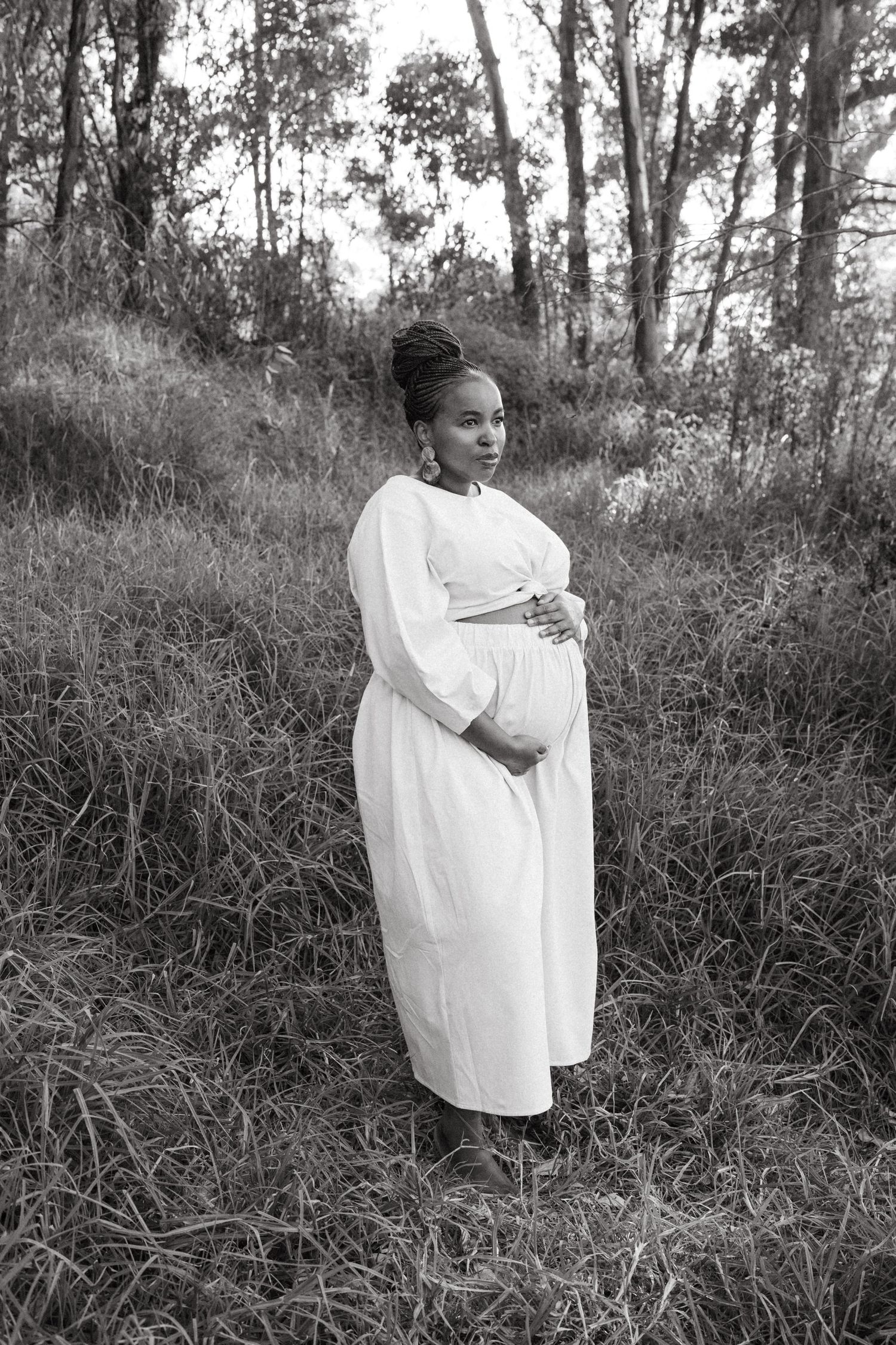 A figure in a flowing white dress stands gracefully in a natural outdoor setting with tall grass and trees.
