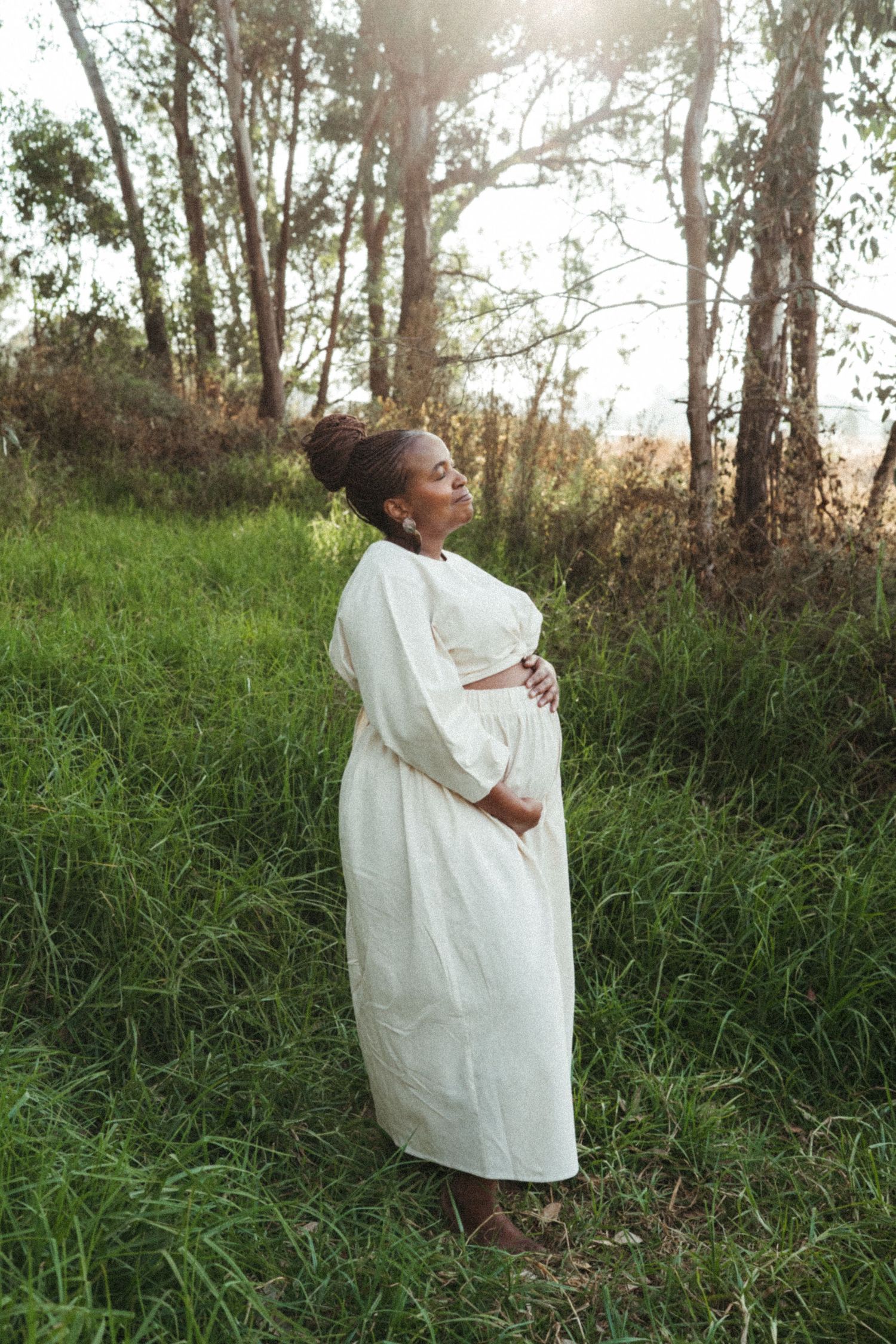Sunlit outdoor scene with a figure wearing white standing among tall grass and trees.
