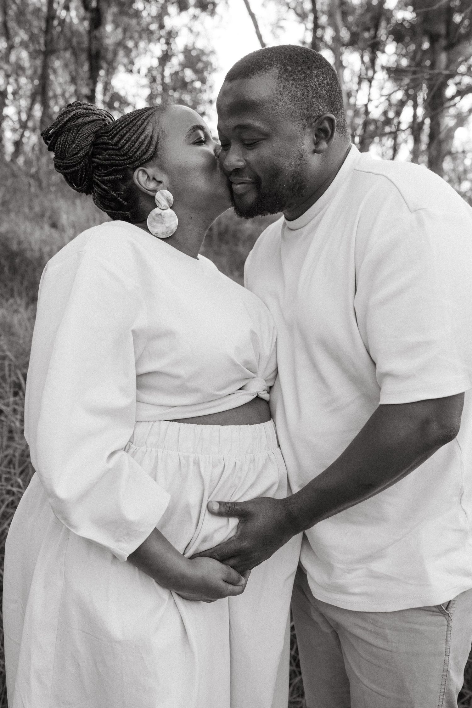 Intimate couple's portrait in white attire embracing outdoors in a wooded setting.
