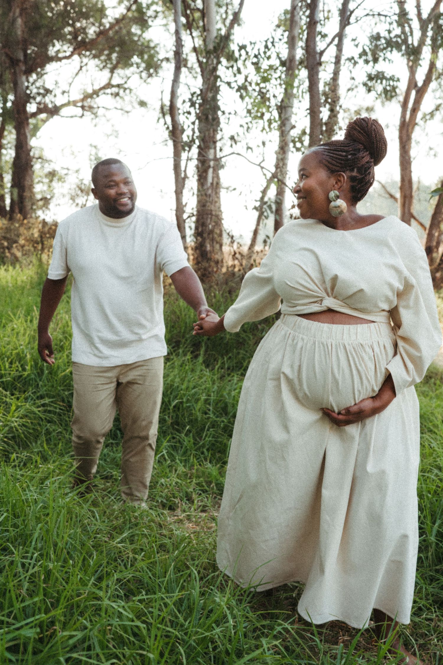 Couple in coordinated white outfits embracing in a sun-dappled wooded area.