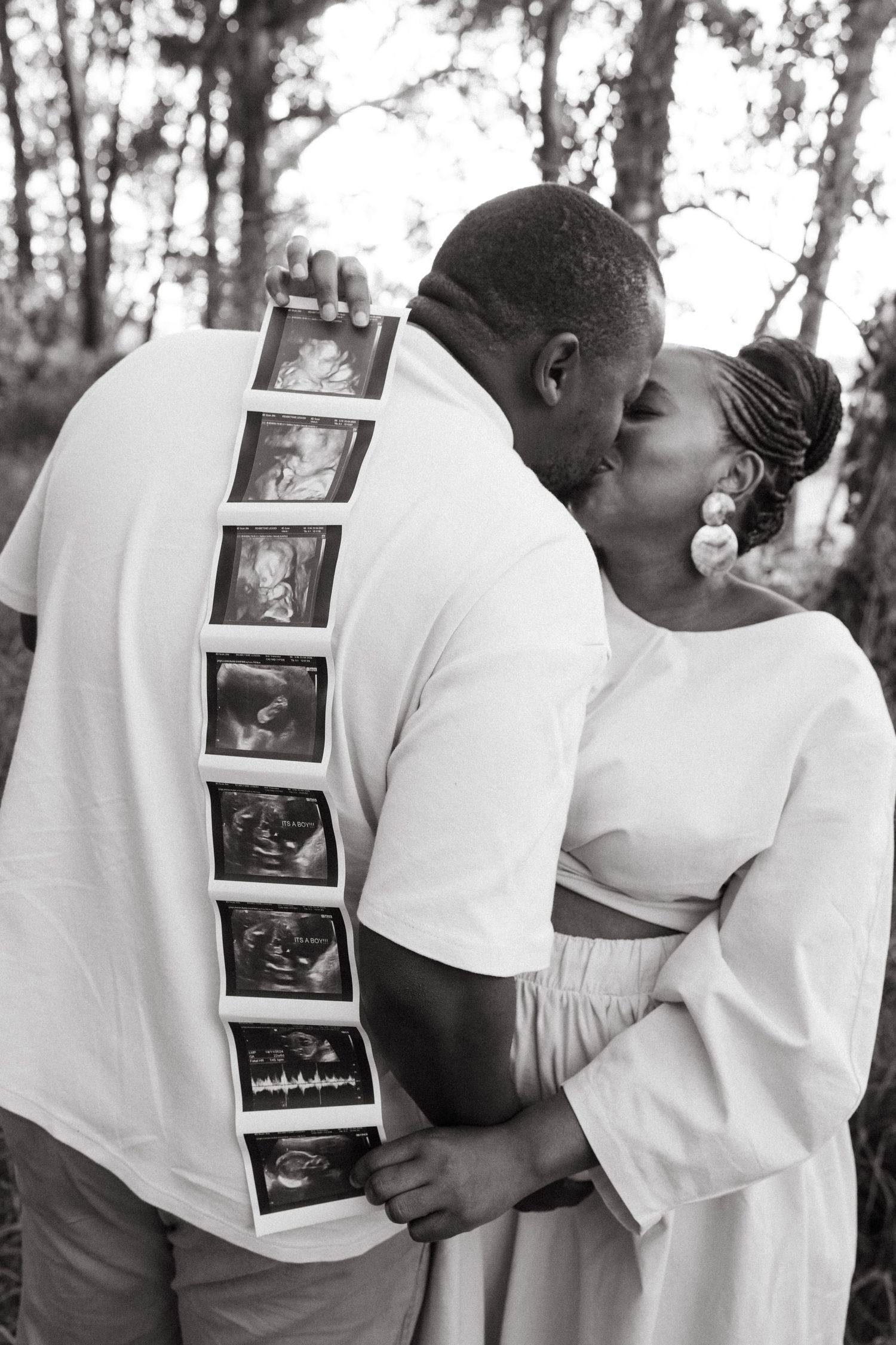 Couple sharing intimate moment while holding ultrasound photos in outdoor setting.