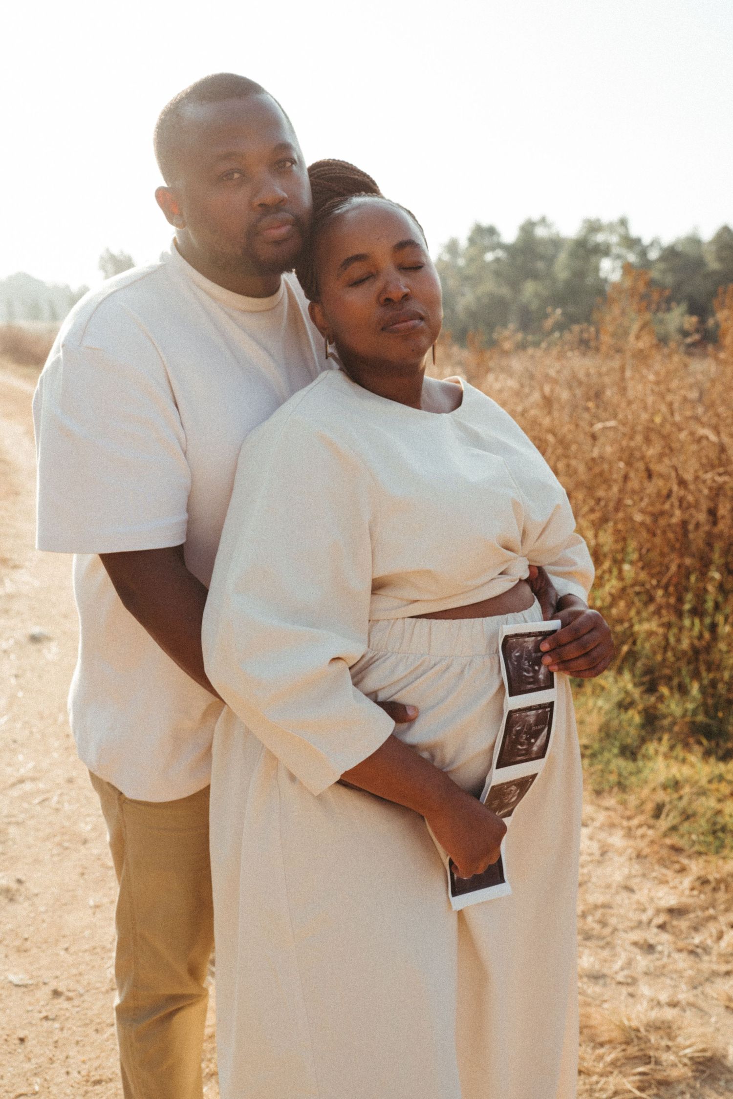 Couple in white clothing embracing while holding sonogram photos in golden sunlight.