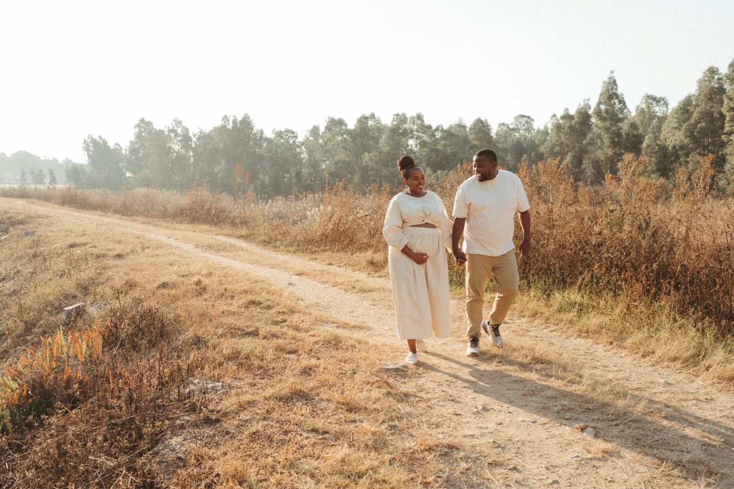Couple walking together on a sunlit dirt path through golden fields.