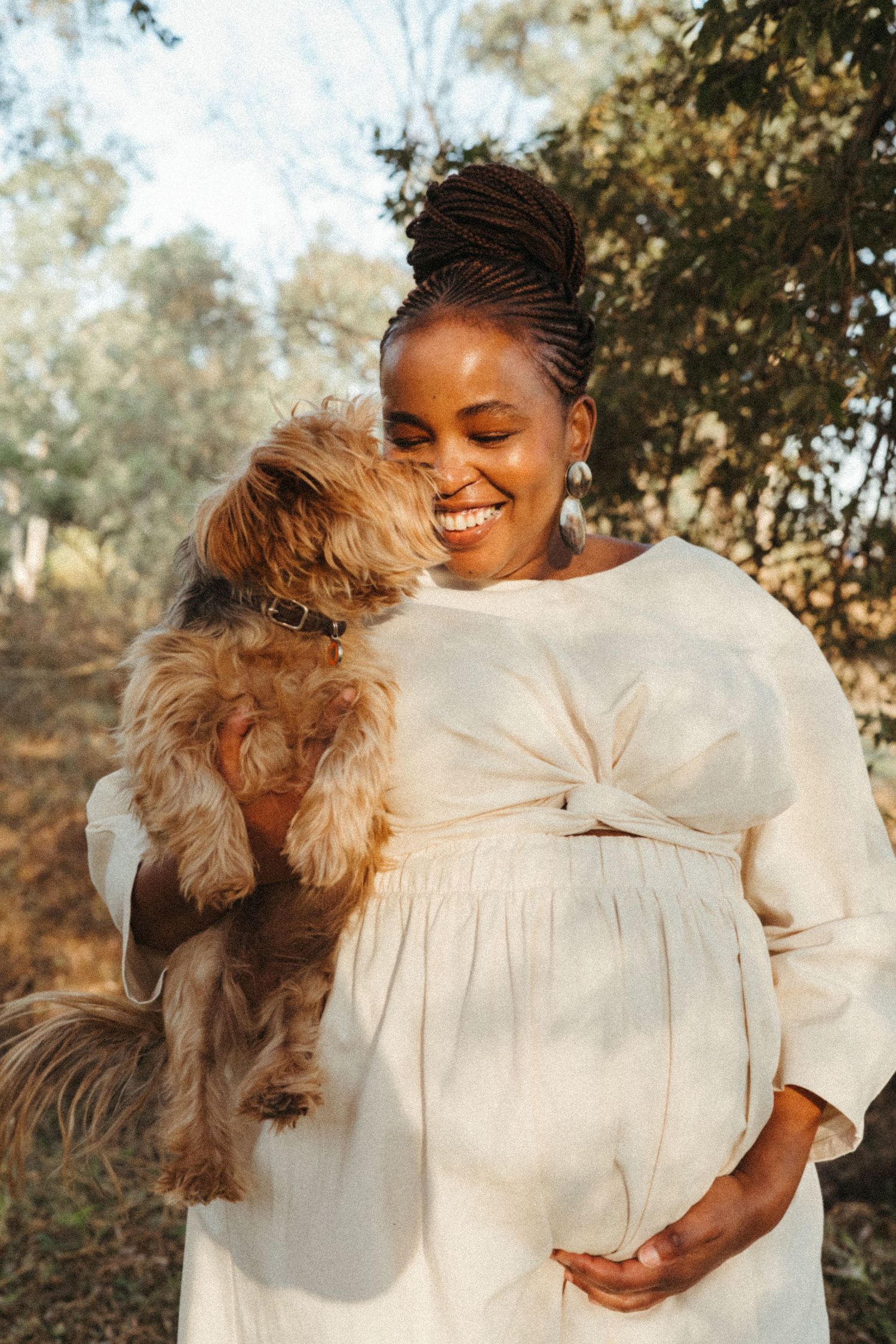 Expecting mother in white dress holding small brown dog while smiling outdoors.