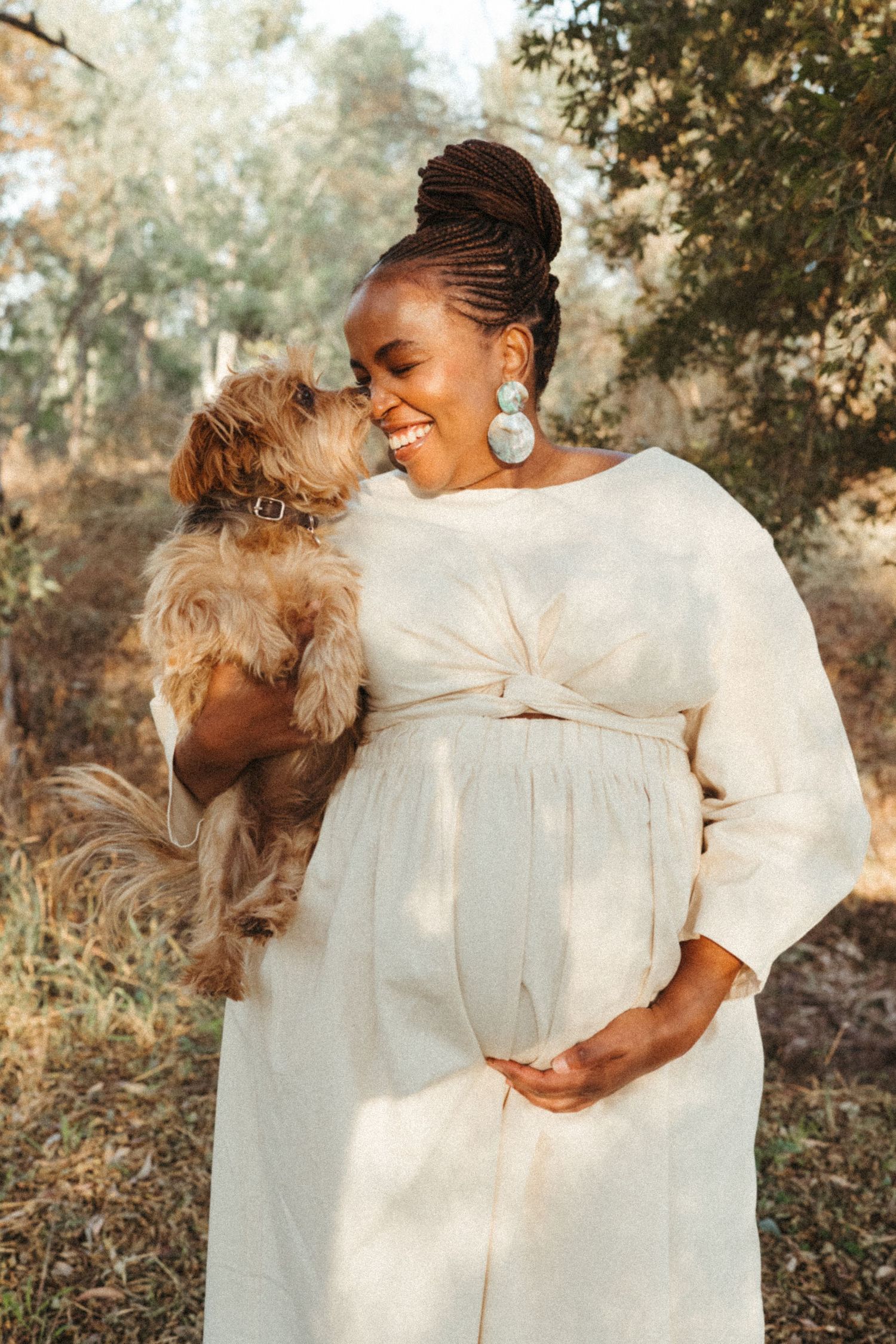 Pregnant woman in white dress shares joyful moment with small brown dog outdoors.