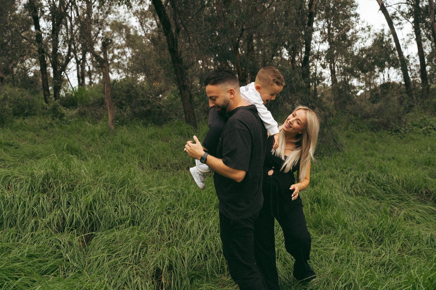 Two people in black clothing embrace while standing in tall grass with trees in the background.