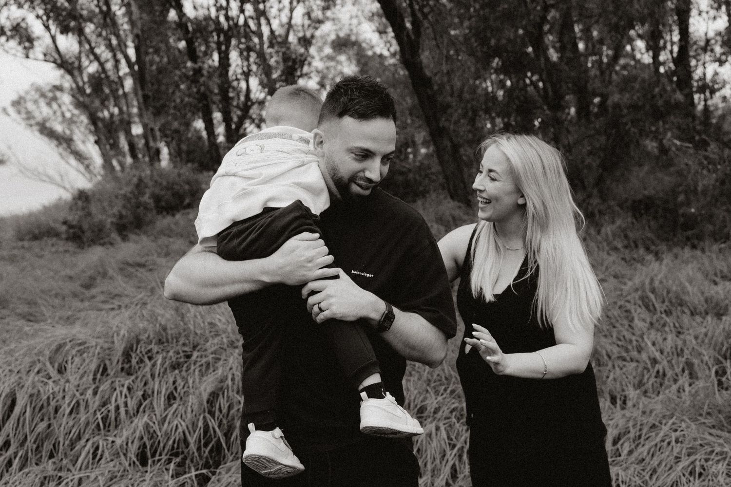 Two people share an intimate moment while standing in tall grass with trees behind them.
