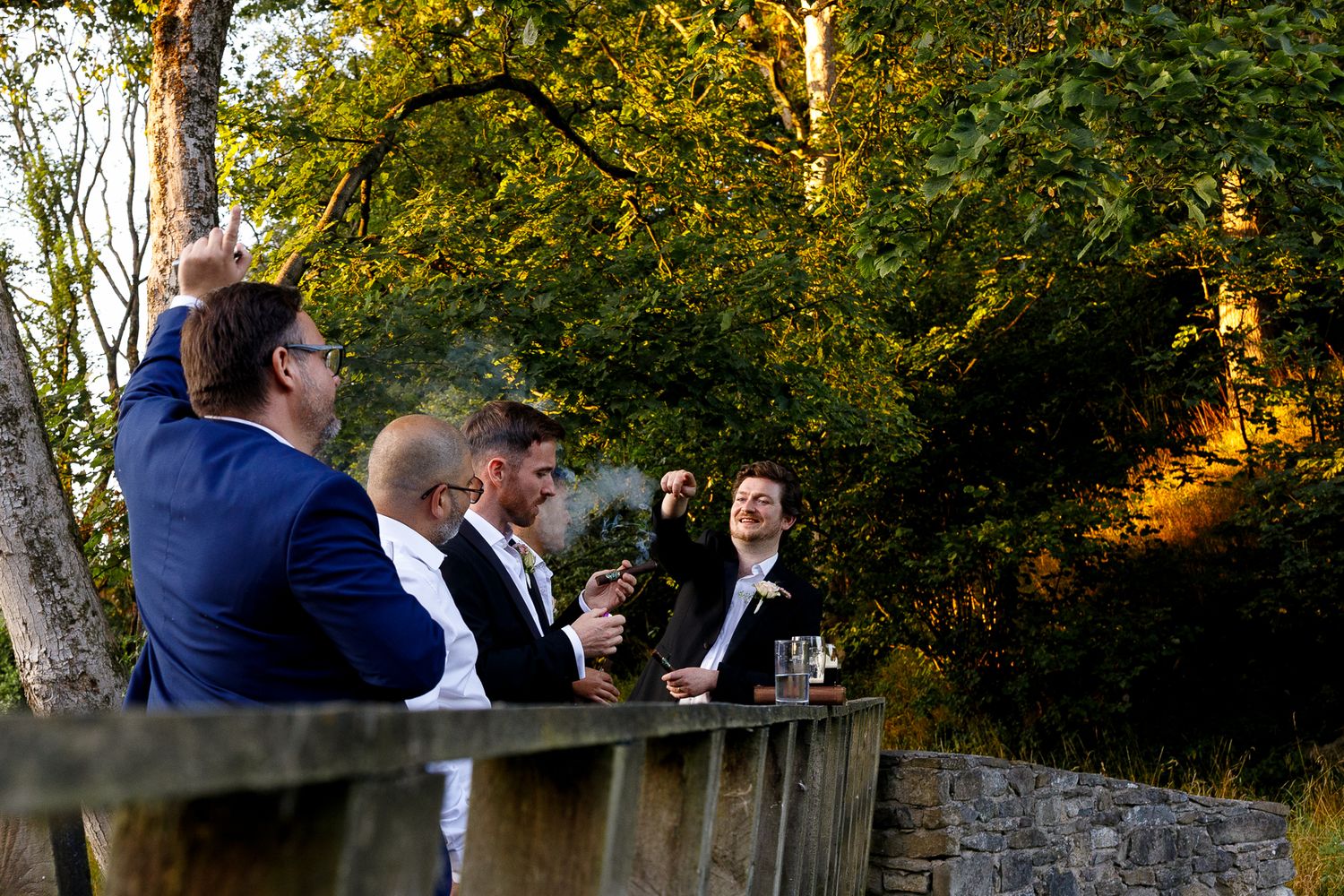 Wedding guests enjoy cocktail hour outdoors at sunset near a stone wall surrounded by autumn foliage.