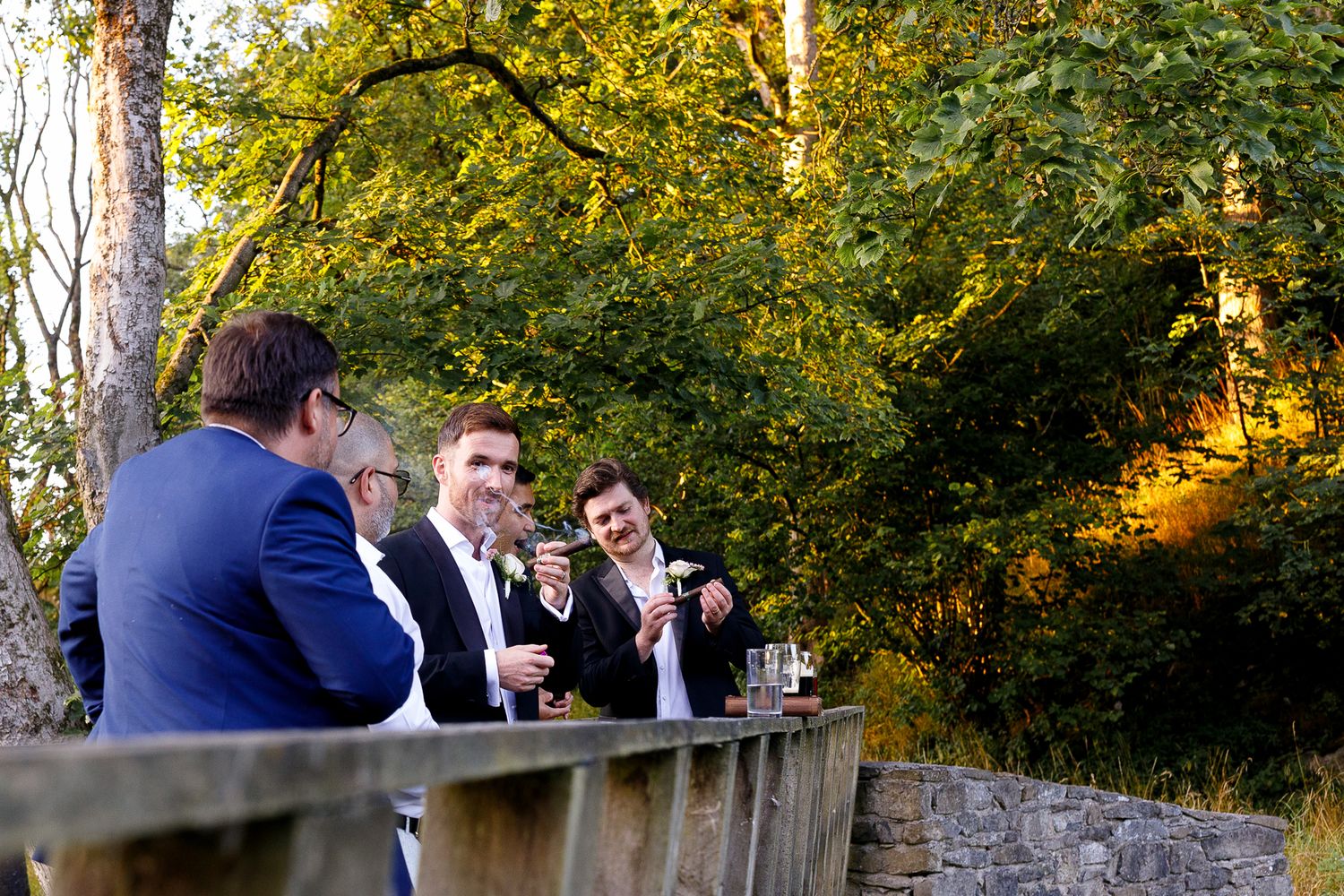 Wedding guests enjoy drinks and conversation along a rustic stone wall during an outdoor reception at sunset.
