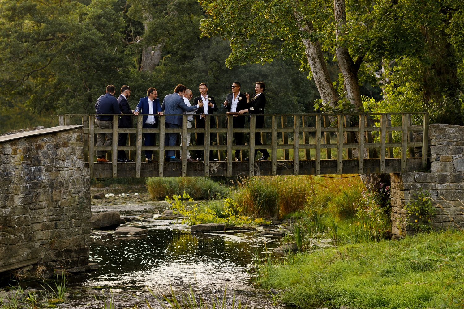 A serene stone bridge spans over a tranquil stream surrounded by lush greenery and trees in a peaceful countryside setting.