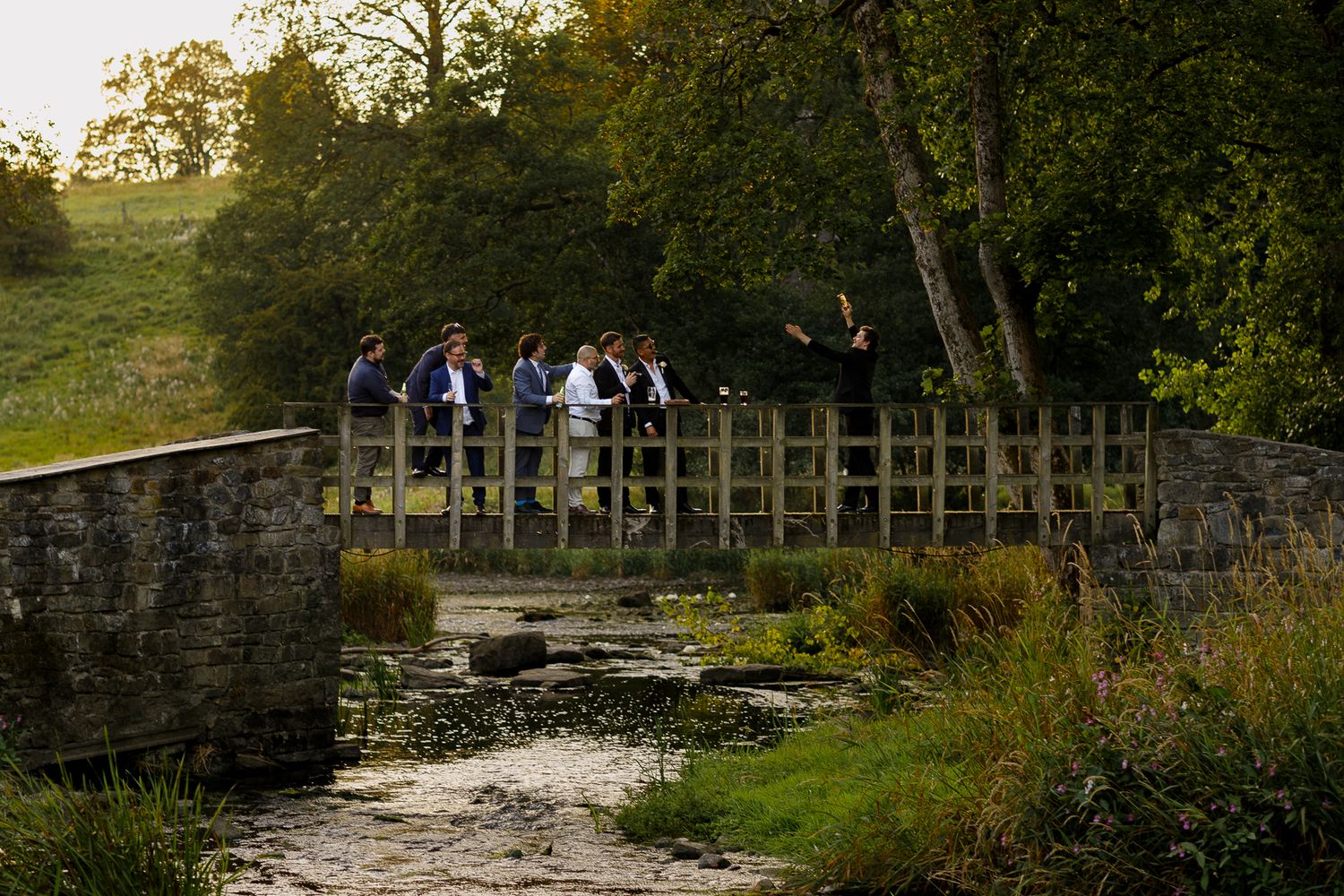 A group of people stand on a stone bridge over a stream at sunset in a scenic rural setting.
