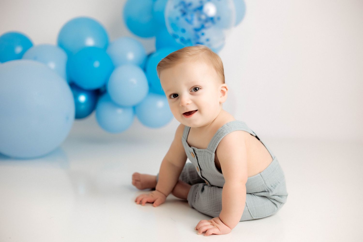 Baby in gray romper sits and plays with blue balloons during first birthday cake smash photo session.