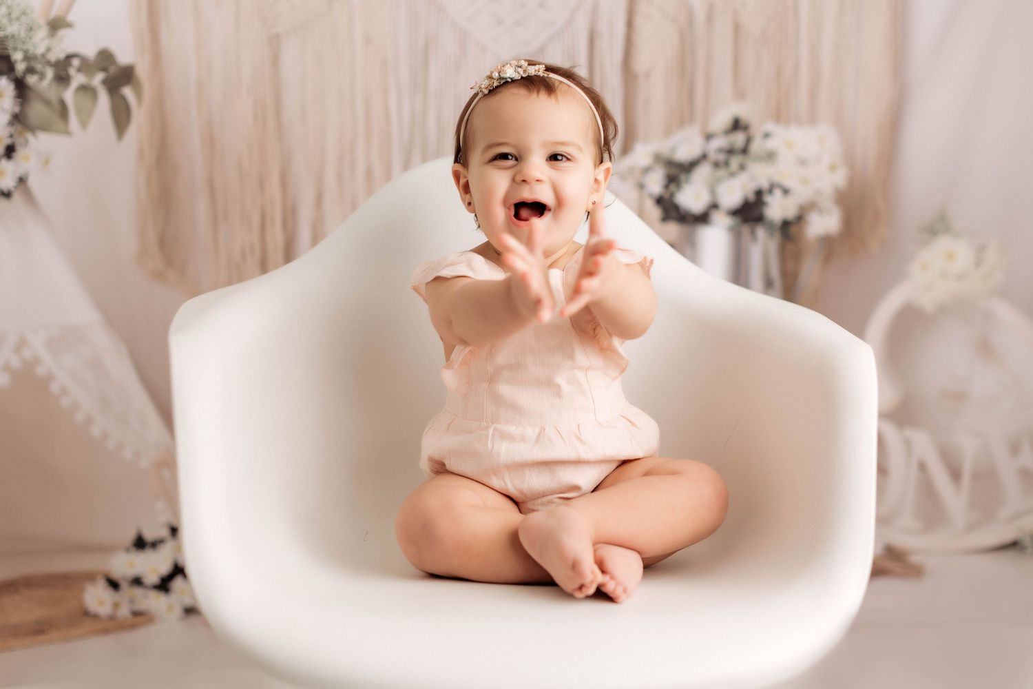 A baby in a white dress sits on a curved modern chair during a professional studio photo session.