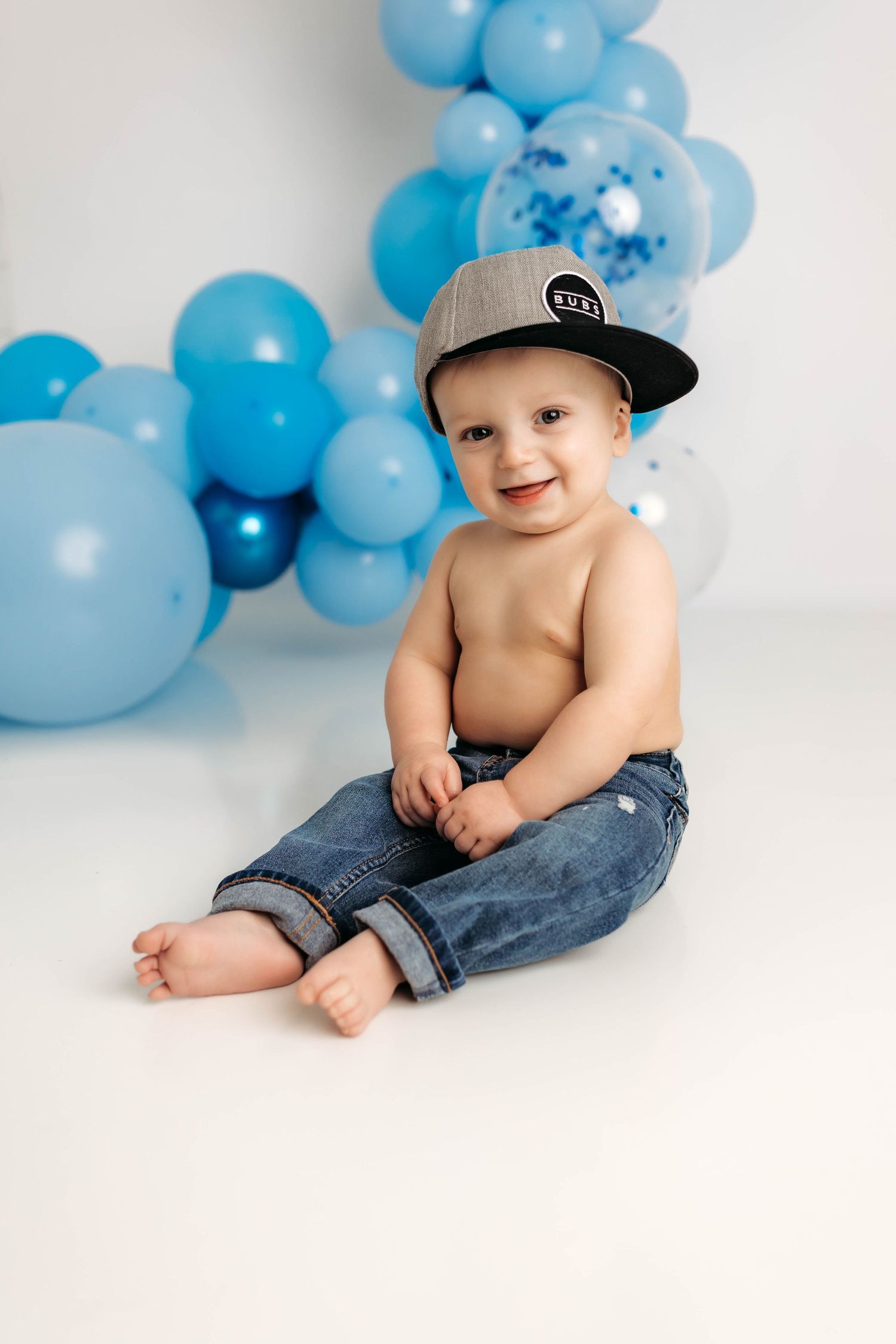 Baby wearing baseball cap and jeans sits against white backdrop with blue balloon decorations.