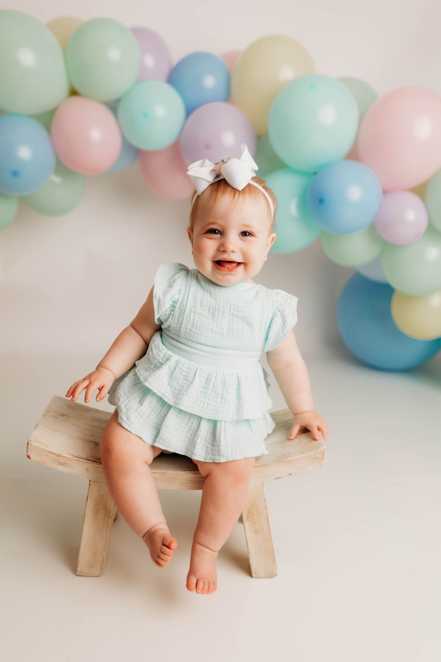 A baby in a mint green dress sits on a wooden stool against a pastel balloon backdrop for a portrait session.