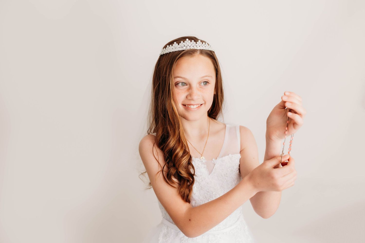 Young dancer in white dress and tiara holds a ballet shoe against light background.
