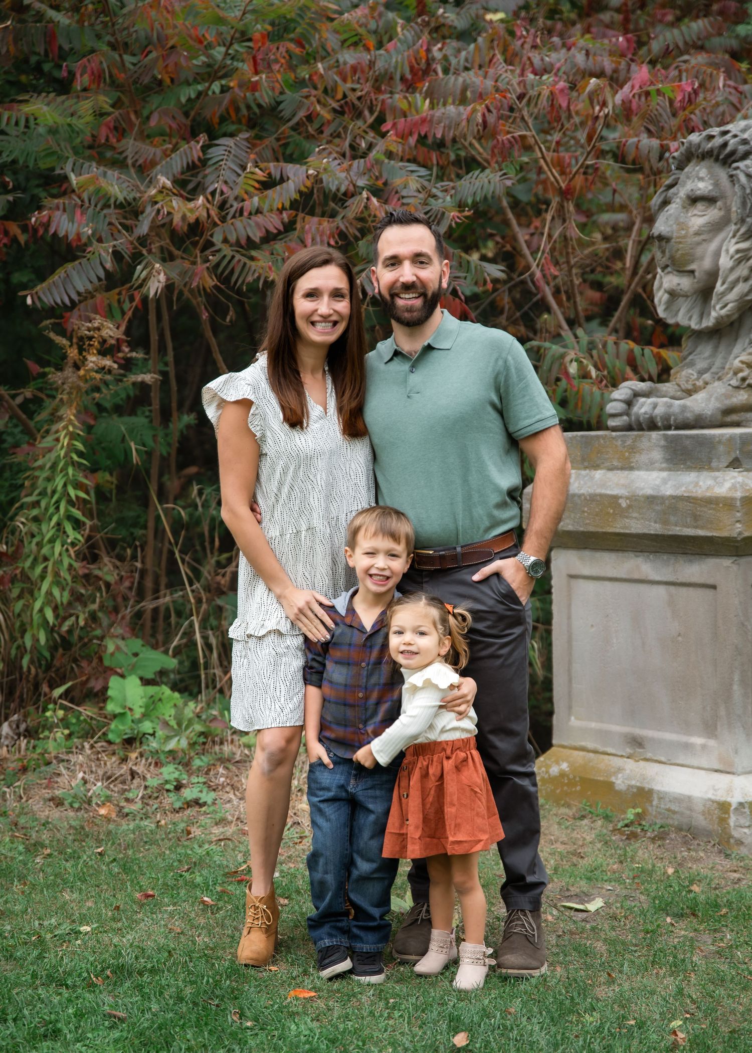 A family poses together outdoors in front of autumn foliage and a stone sculpture during a portrait session.