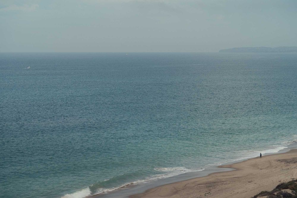 A panoramic view of a sandy beach stretches along the coastline with turquoise ocean waters and distant cliffs.