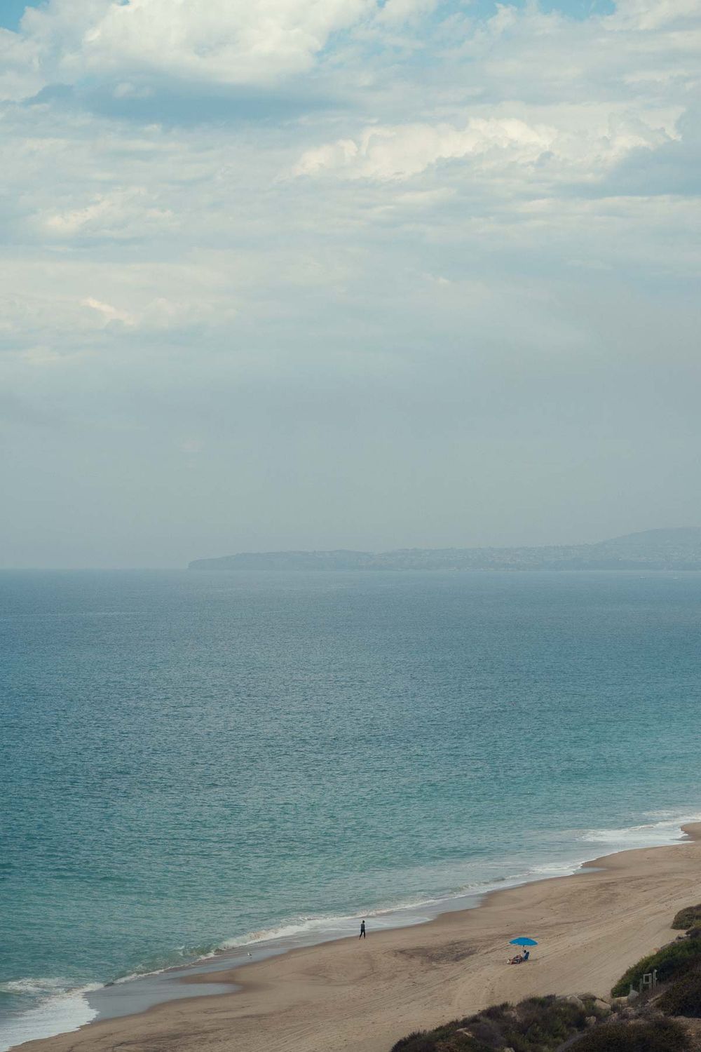 A panoramic view of a serene beach with turquoise waters, distant mountains, and a lone blue umbrella on the sand.