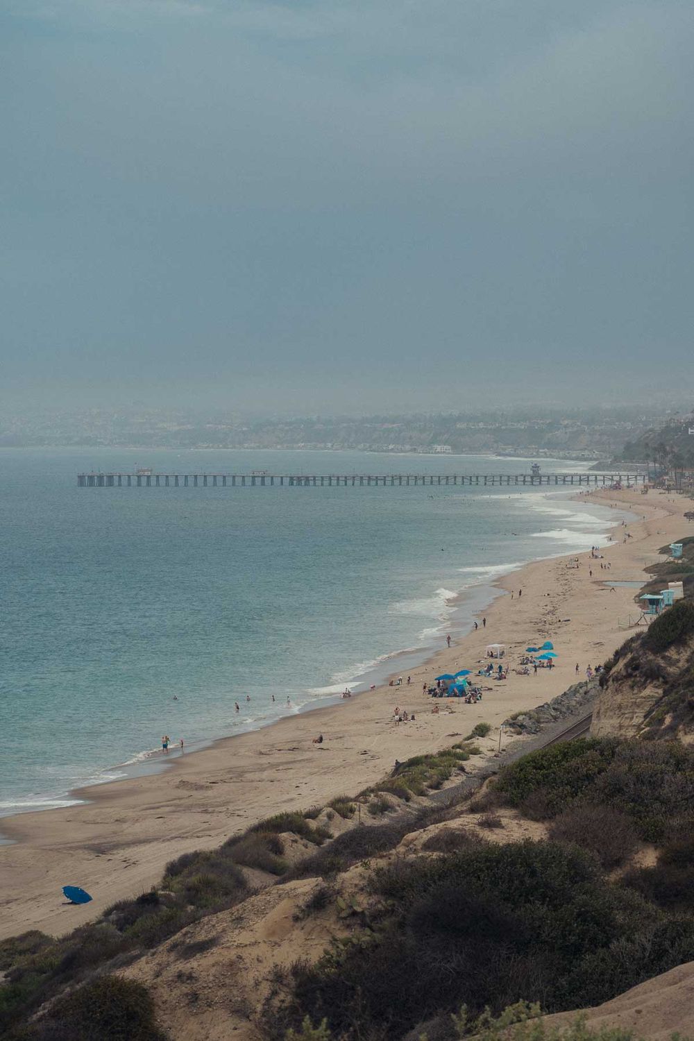 Aerial view of a sandy beach with scattered beachgoers and a long wooden pier stretching into the misty ocean.