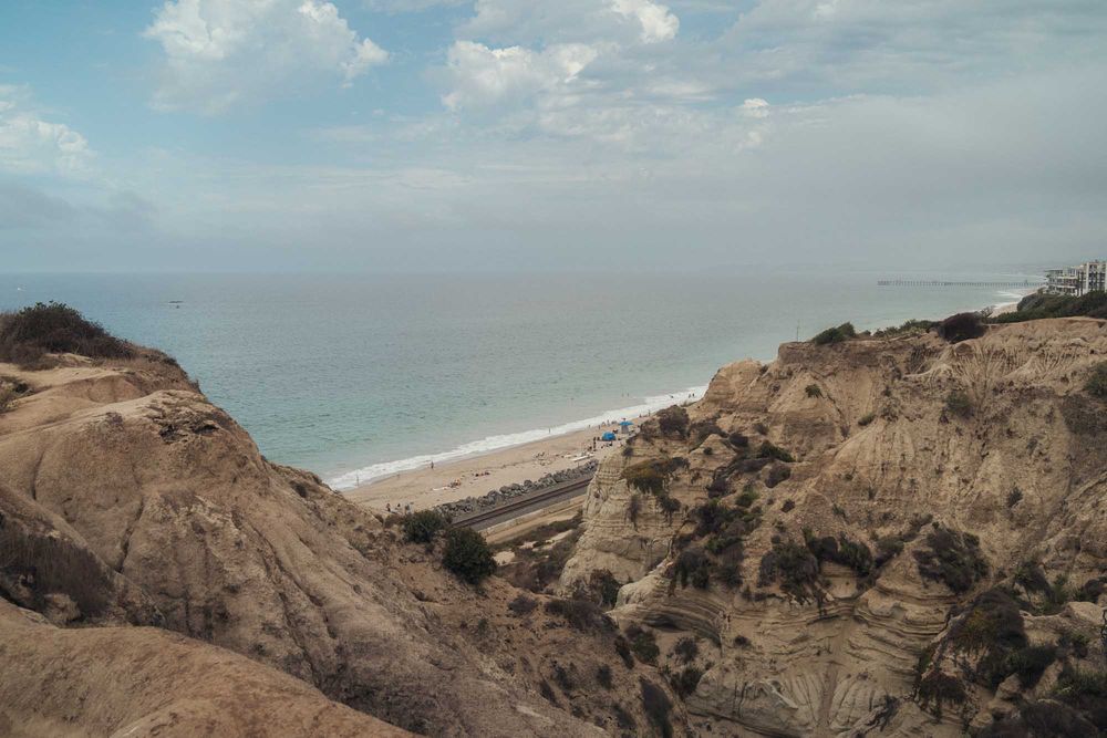 Aerial view of rugged coastal cliffs overlooking a sandy beach and blue ocean under cloudy skies.
