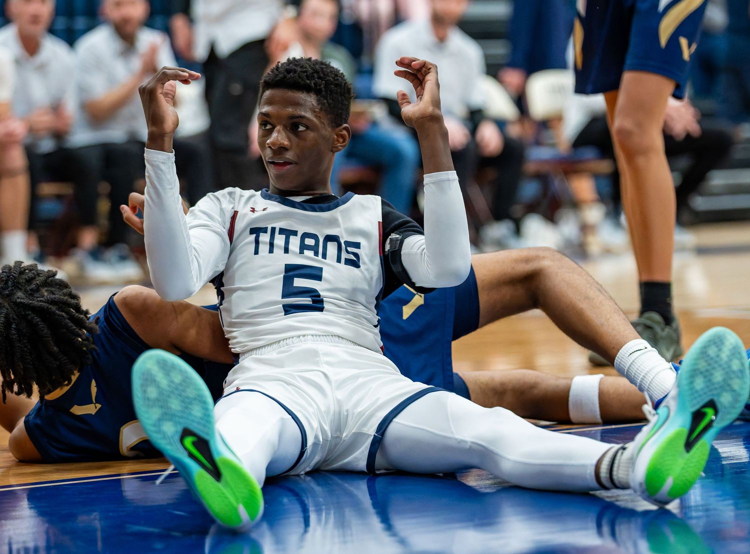 Basketball player in white Titans jersey number 5 sits on court gesturing with both hands raised during game.