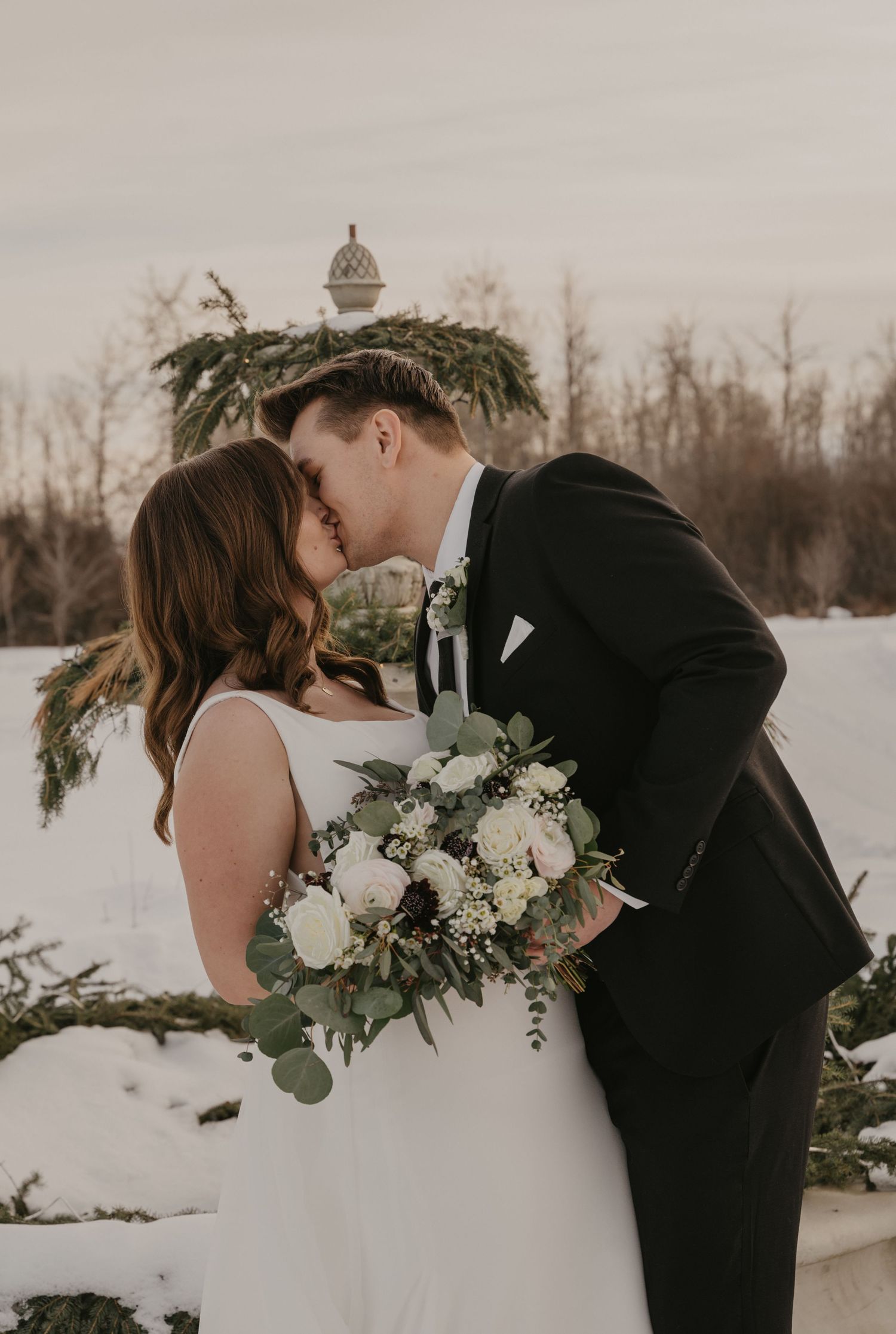 A romantic winter wedding photoshoot shows a couple in black and white attire embracing in the snow.