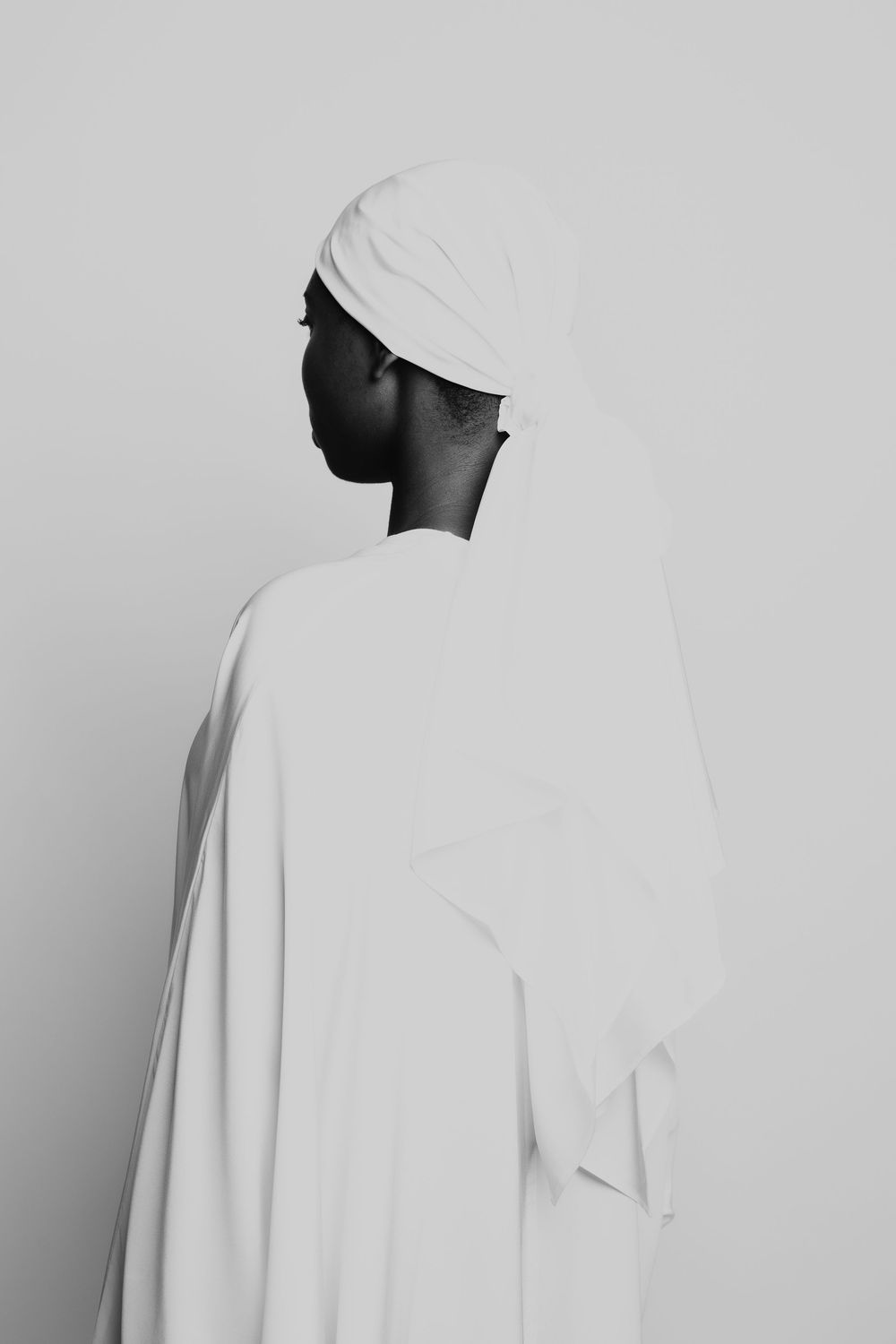 Minimalist black and white portrait showing silhouette in white head wrap and flowing garment against light background.