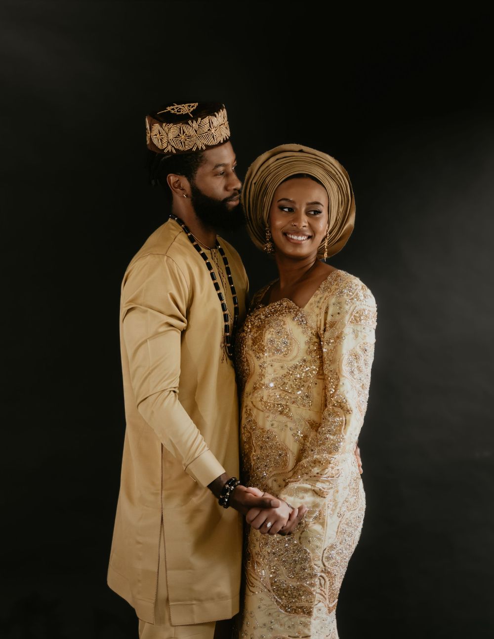 Couple in traditional West African attire share intimate moment during photo session against dark backdrop.