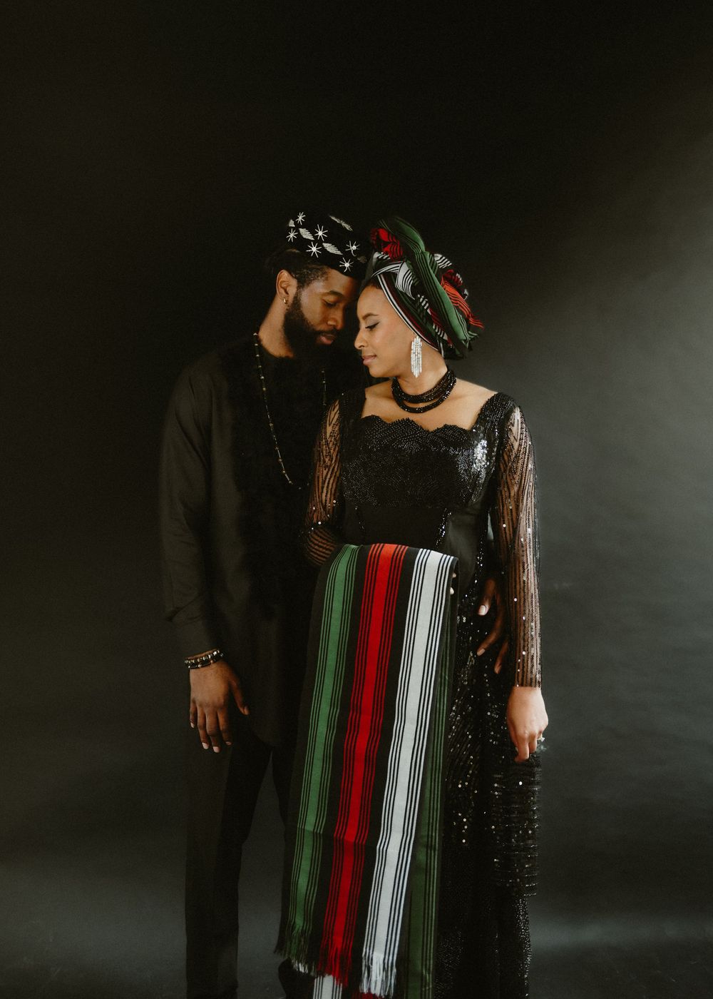 A couple in traditional Nigerian attire poses with red and white striped fabric against a dark background.