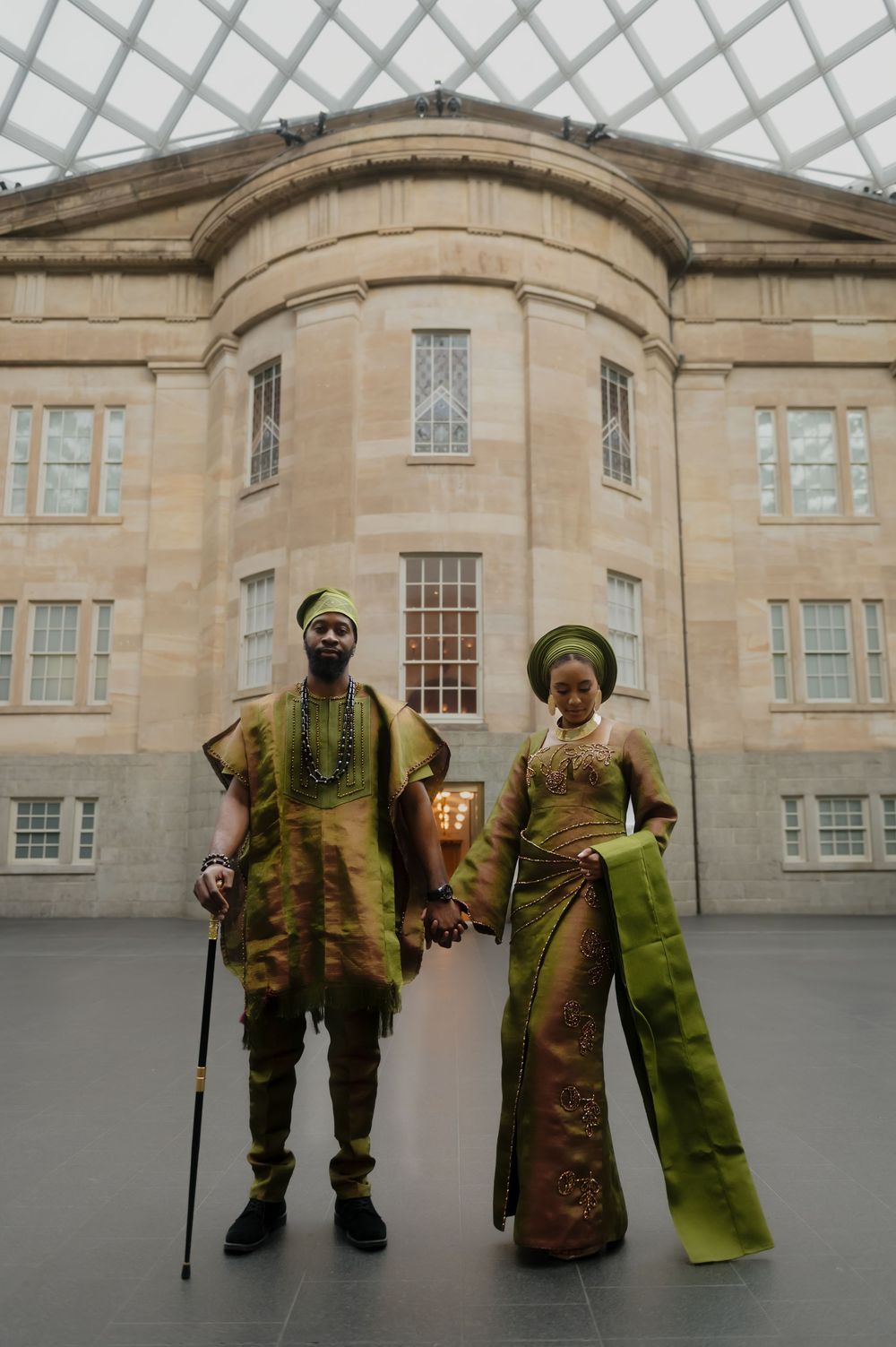 Two people in green and brown traditional African attire pose together on stone courtyard.