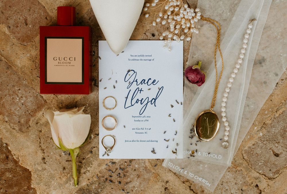 Wedding details flatlay with invitation, rings, flowers, and burgundy accents on textured beige background.