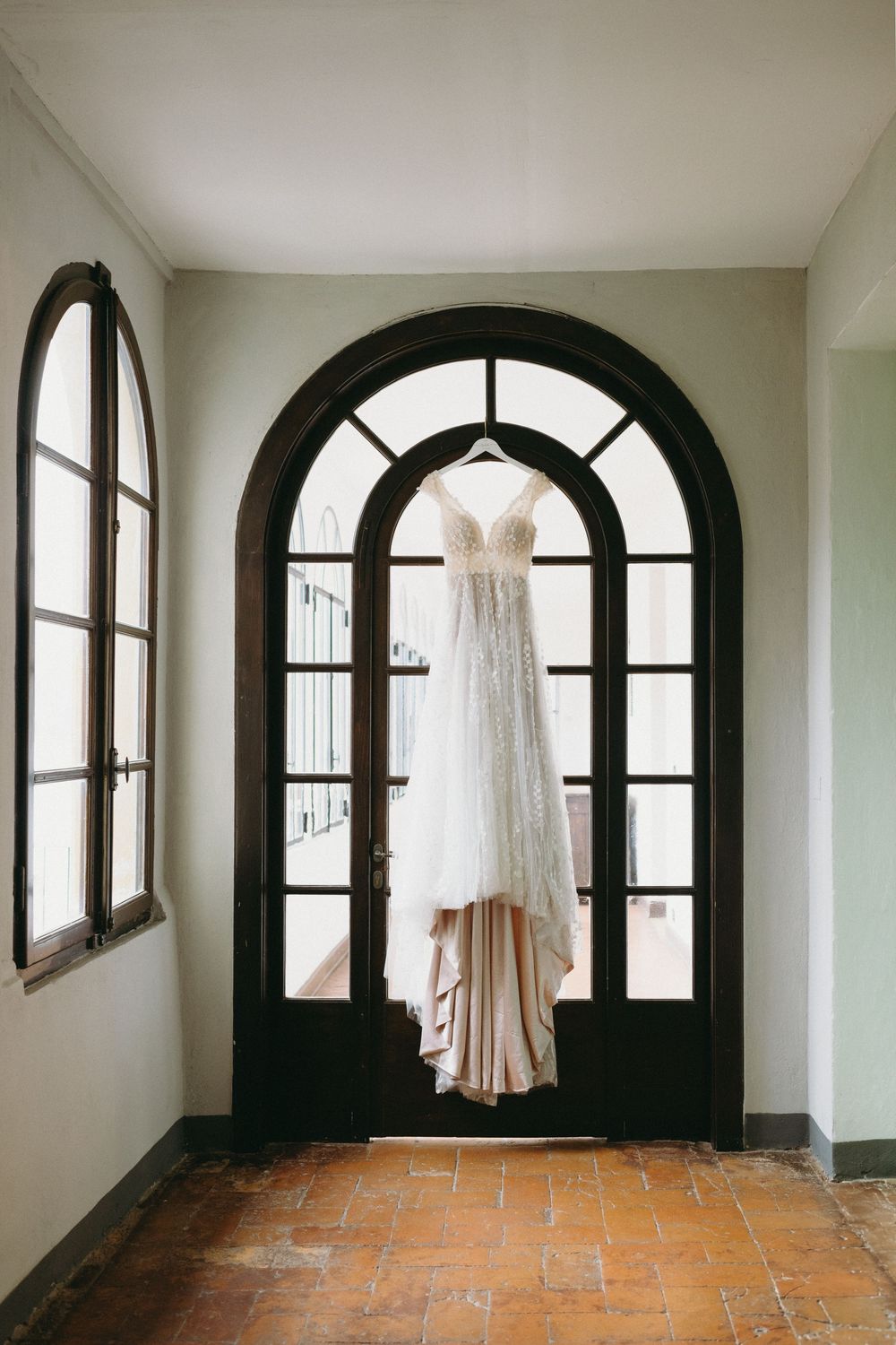 A white wedding dress hangs elegantly against arched black windows in an empty room with terracotta floors.